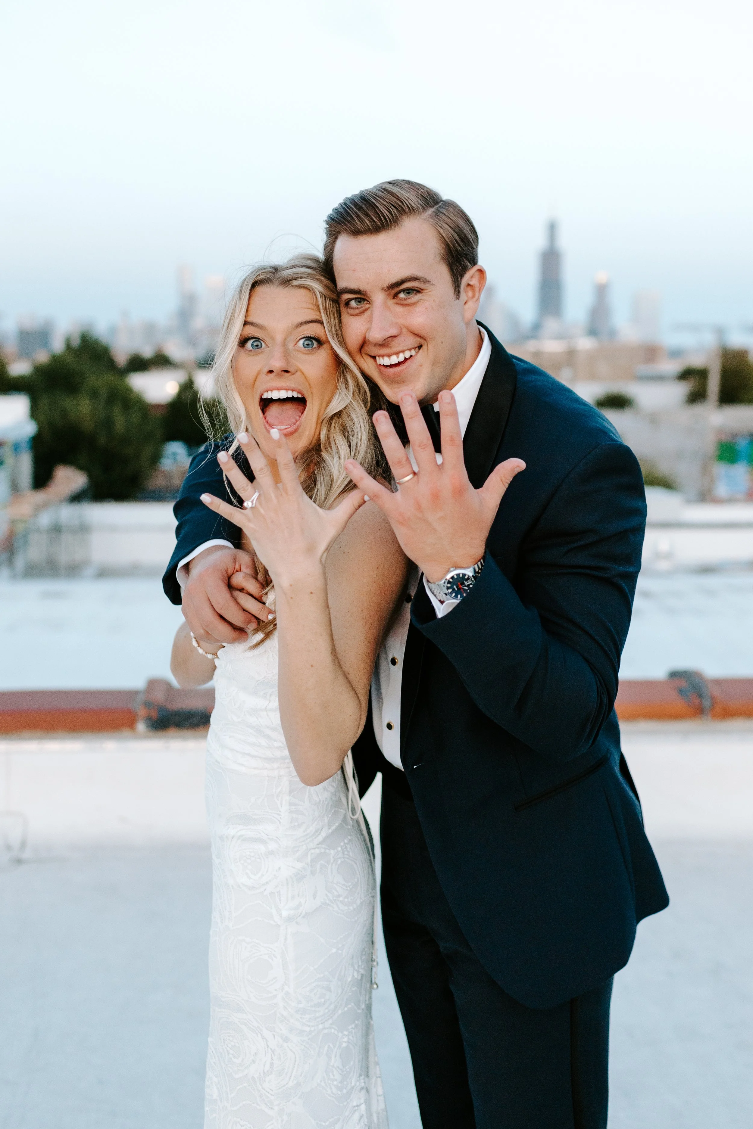 A newlywed couple celebrating with the woman showing off her engagement and wedding rings on a rooftop with a city skyline in the background.