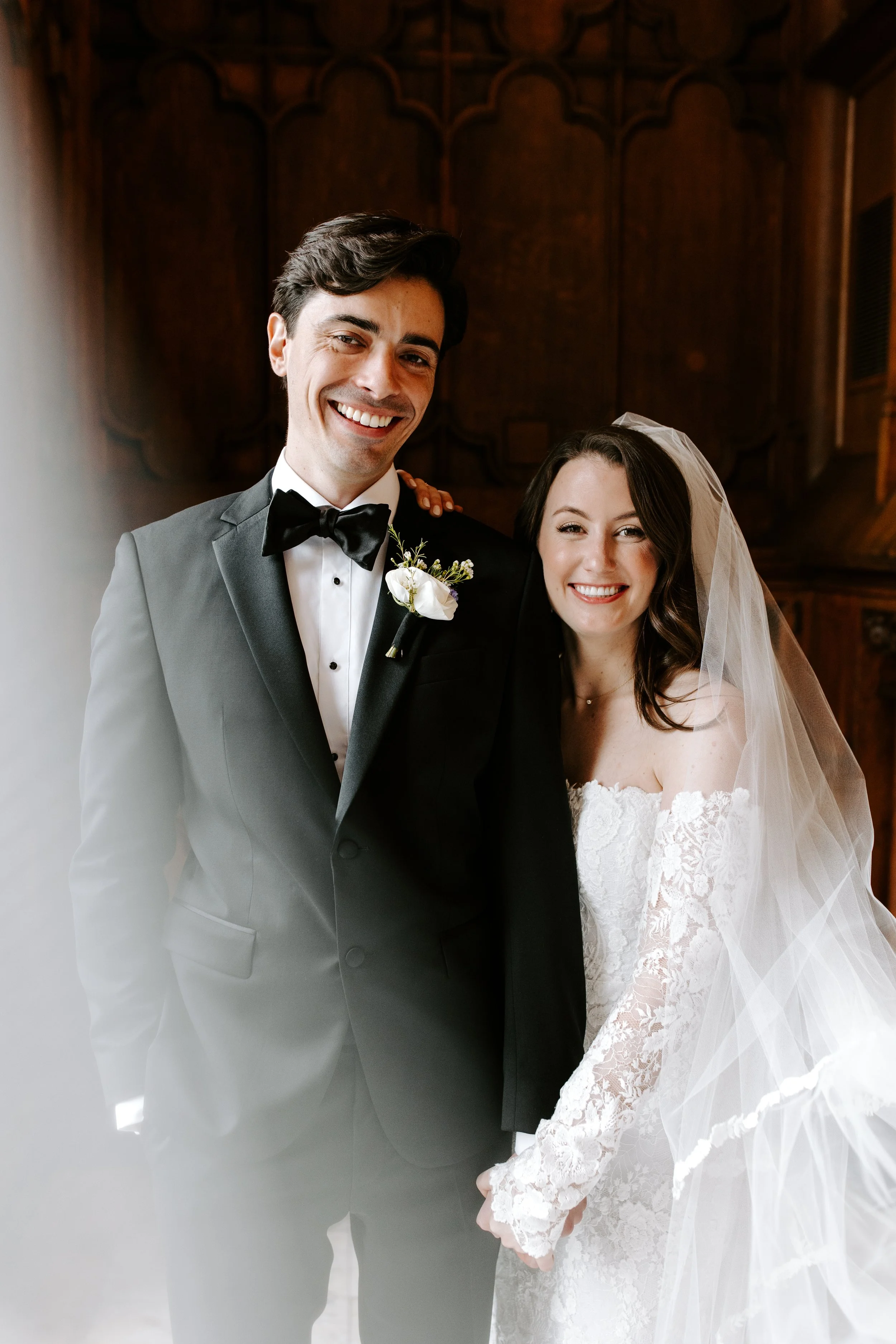 A bride and groom smiling together in a church. The groom wears a black tuxedo with a bow tie and boutonniere, and the bride wears a lace wedding dress and veil.