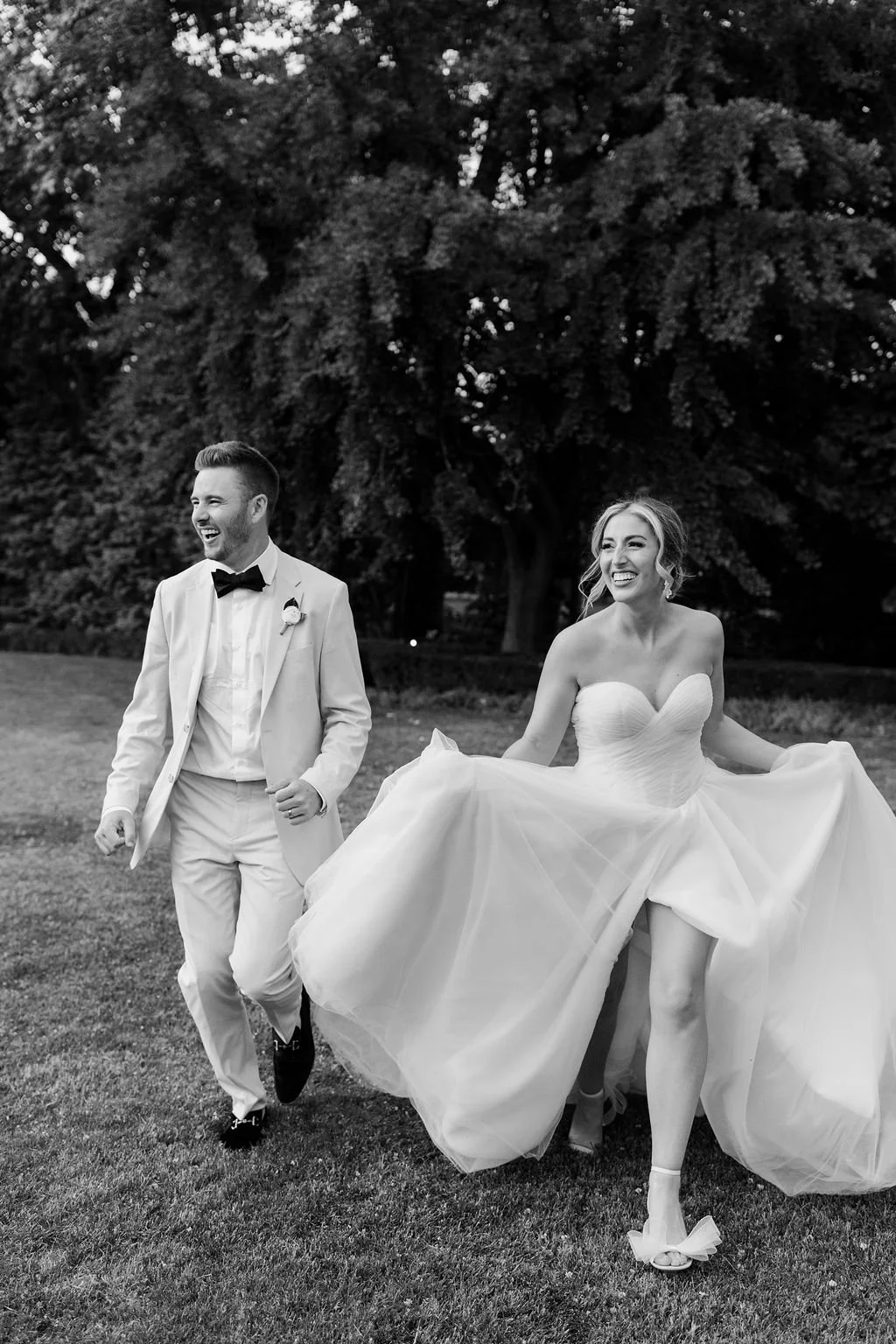 A bride and groom in wedding attire walking outdoors and smiling, with a backdrop of large trees.