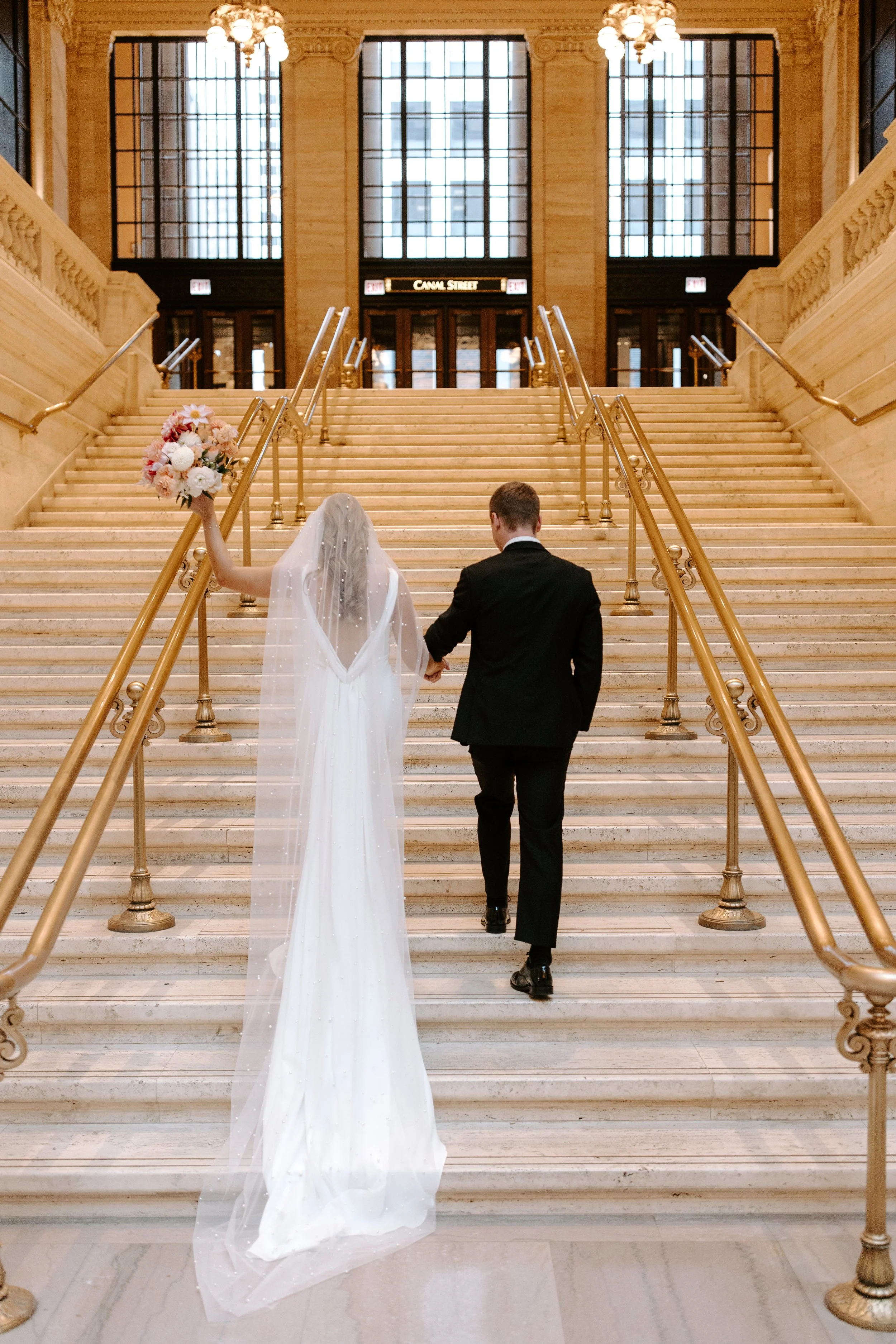 A bride and groom ascending a grand marble staircase inside a building, with the bride holding a bouquet and wearing a white wedding gown and veil, while the groom is dressed in a black suit.