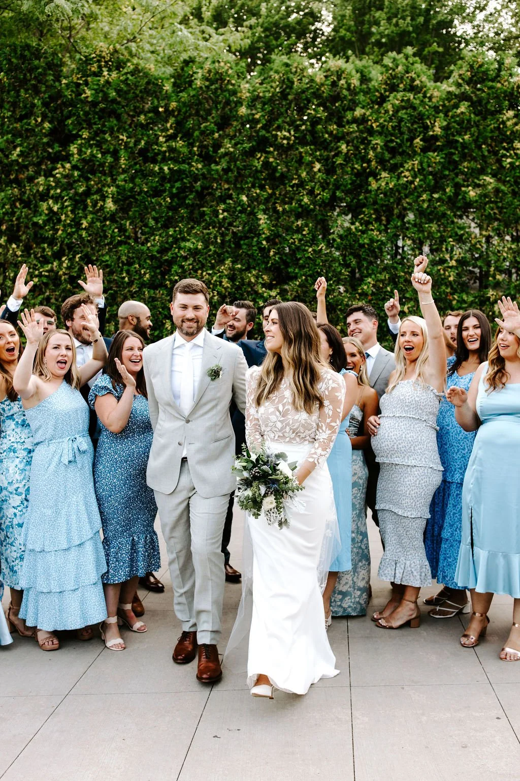 Wedding celebration with bride and groom walking together surrounded by smiling guests, outdoors with green trees in background.
