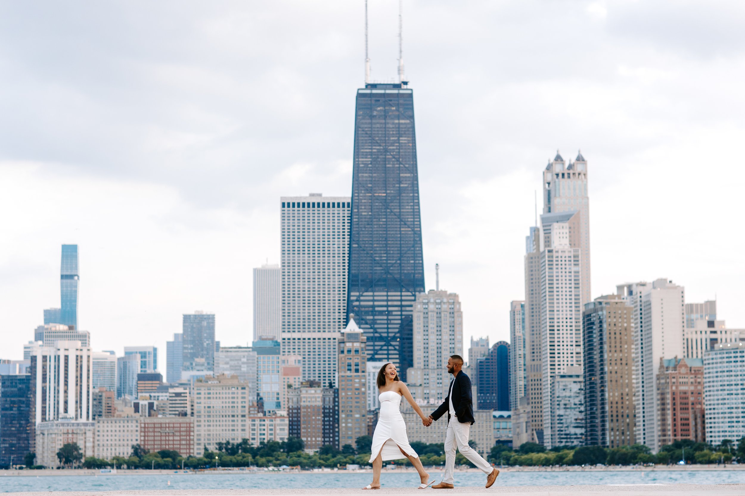 A couple holding hands and walking along a waterfront with the Chicago skyline in the background.