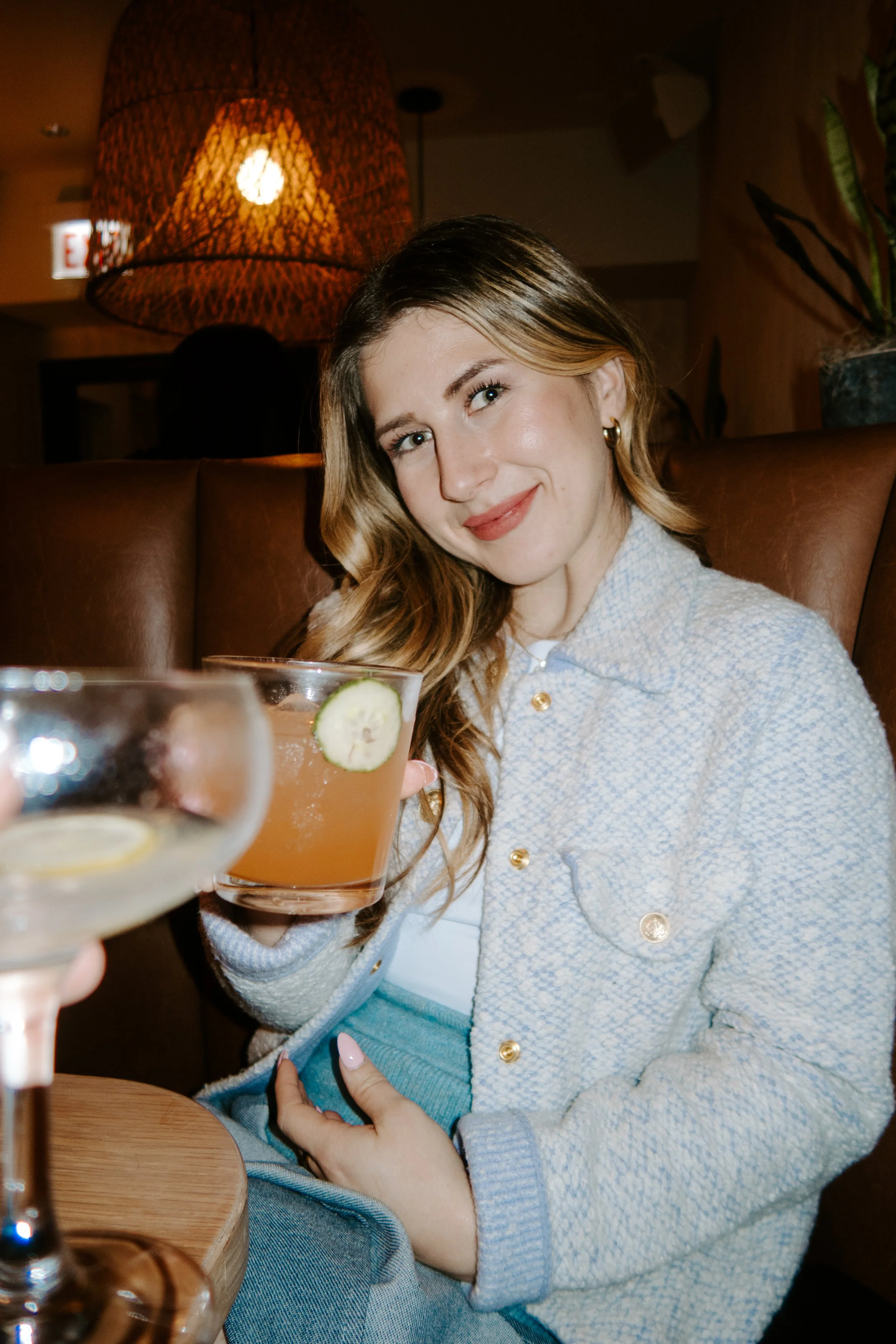A woman smiling, holding a cocktail with a cucumber slice garnish, sitting in a cozy restaurant with warm lighting and a wicker lampshade.