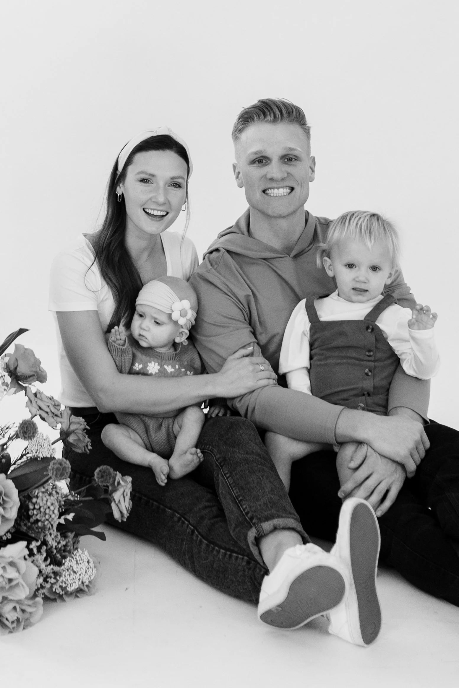 A black-and-white photo of a smiling family of four. The mother and father are sitting on the floor with two young children, a girl and a boy, on their laps. The mother has long dark hair and is wearing a headband, and the father has short styled hair. The girl is wearing a headband with a flower, and the boy is wearing overalls. There are flowers partially visible on the left side of the image.