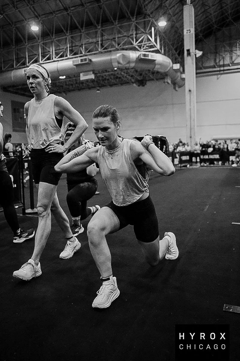 A female athlete in a gym, kneeling on one knee with her hands on her shoulders, wearing athletic clothing and sneakers, with another person standing nearby, all in a black and white photo.
