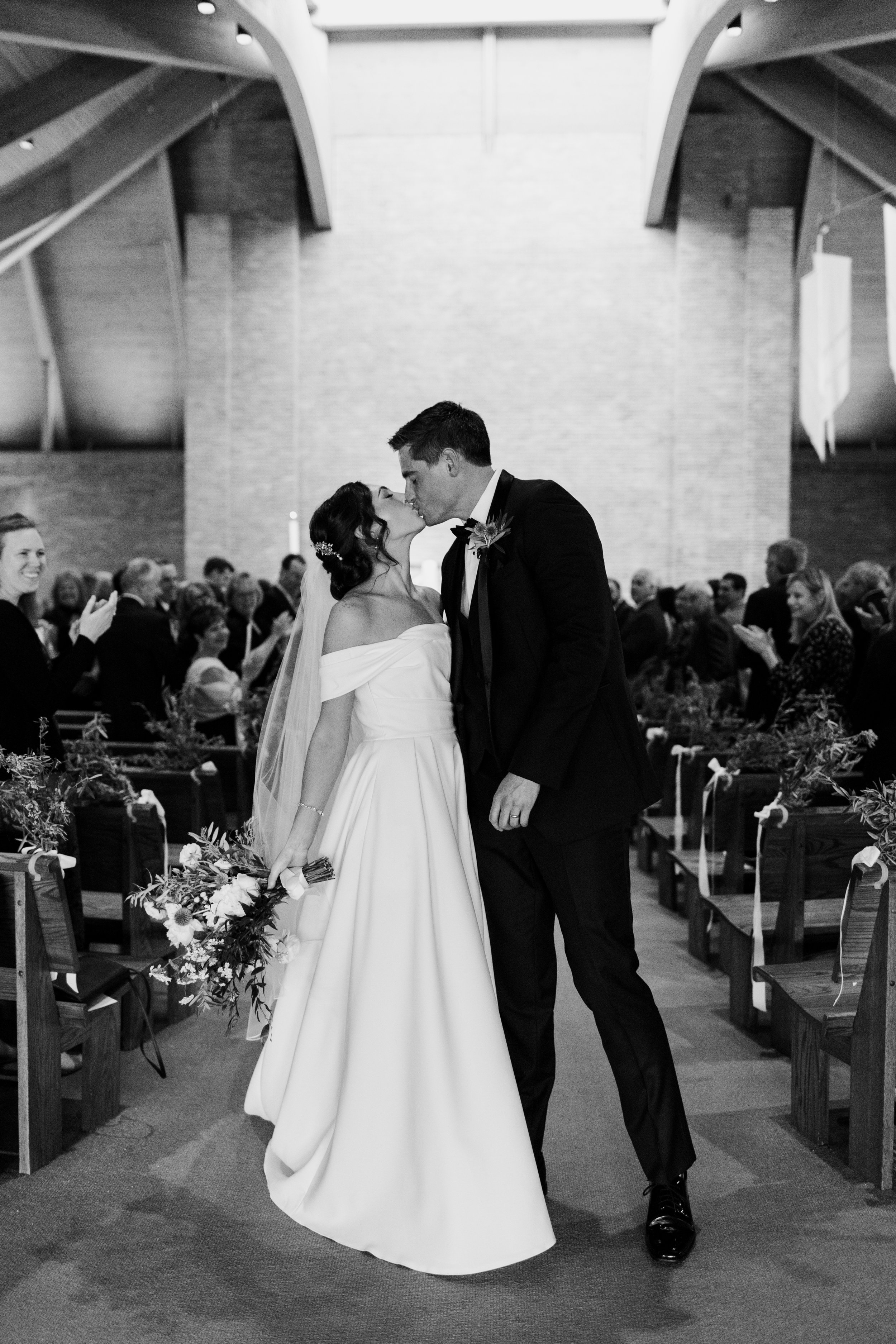 A black and white photo of a bride and groom kissing at their wedding ceremony inside a church, with guests clapping and smiling in the background.