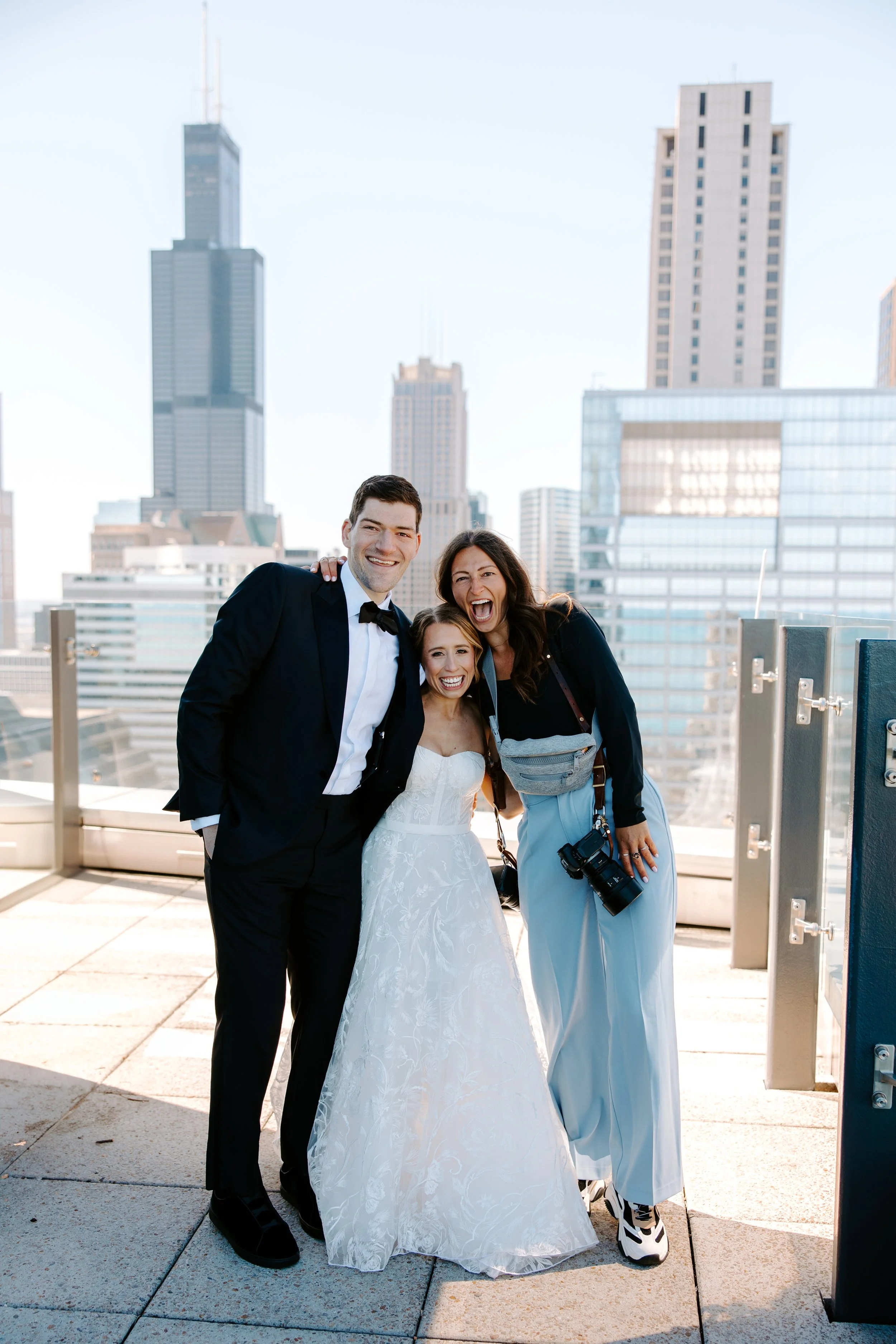 Three people smiling on a rooftop with cityscape background, one in a wedding dress, one in a tuxedo, and one in casual clothes holding a camera.