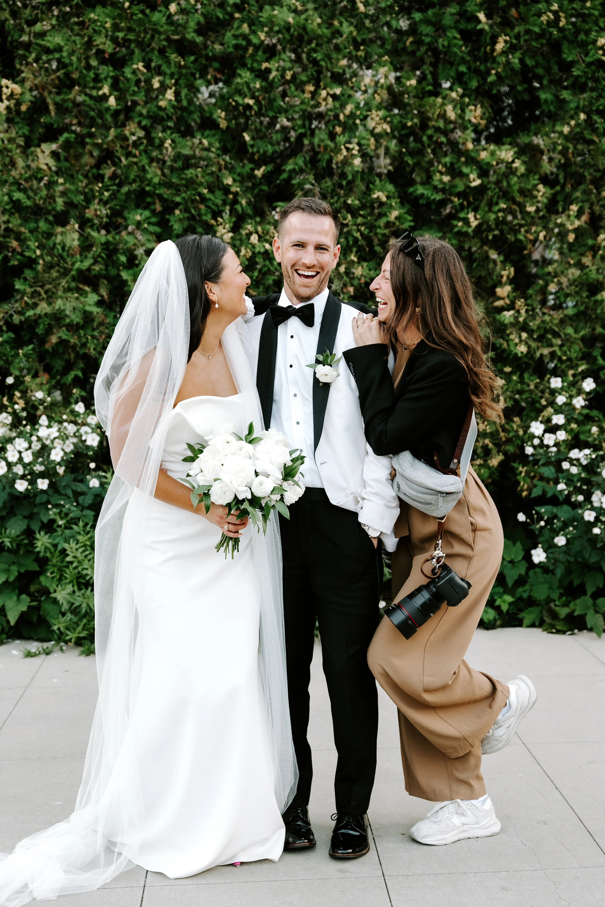 A bride in a white wedding dress holding a bouquet of white flowers, a groom in a tuxedo, and a woman with long brown hair, glasses, tan pants, and a camera, laughing together outdoors in front of a green hedge.