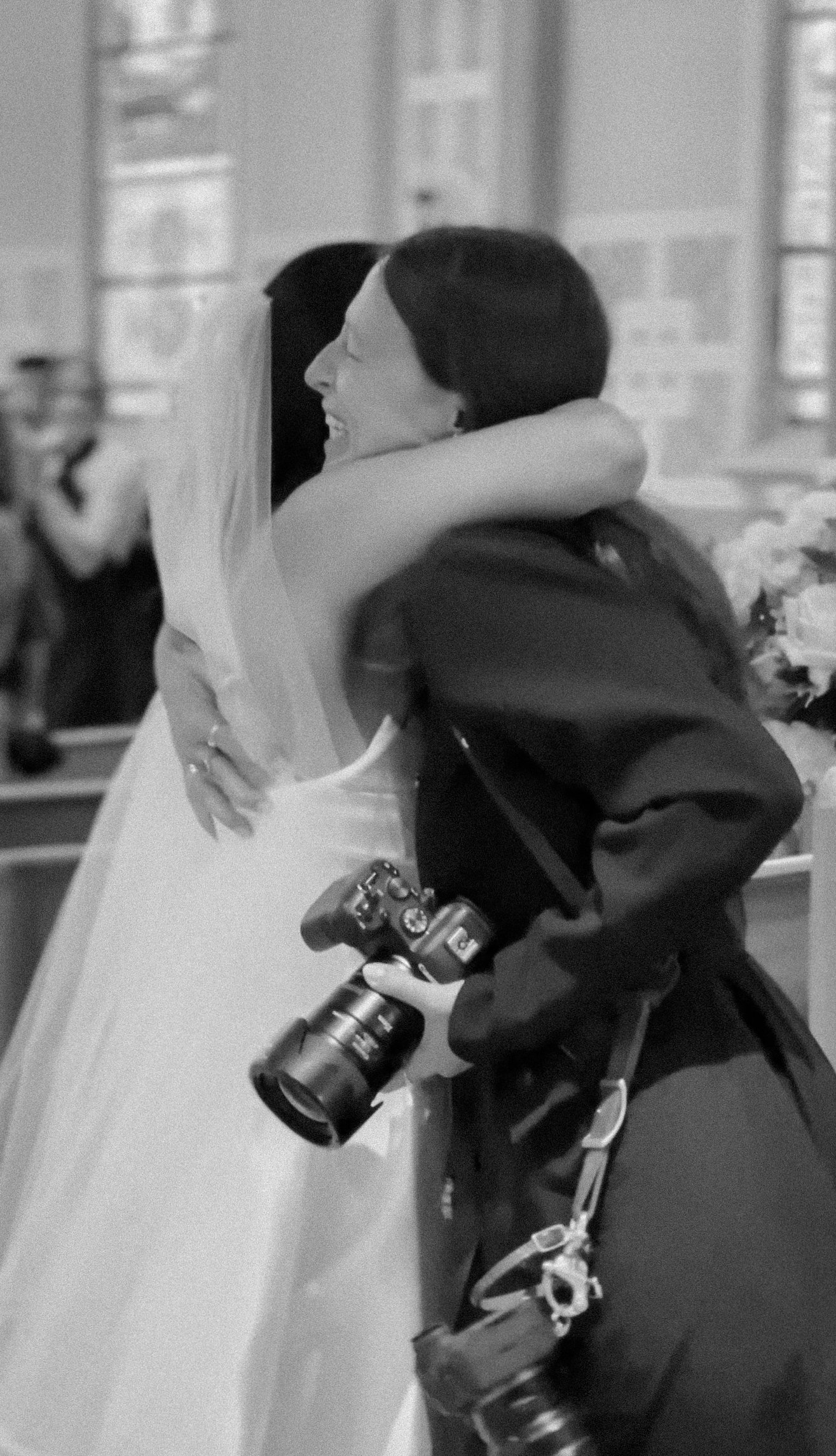 A person with a camera hanging around their neck takes a photo of two women hugging, with one woman in white and the other in dark clothing, in a room with large windows.