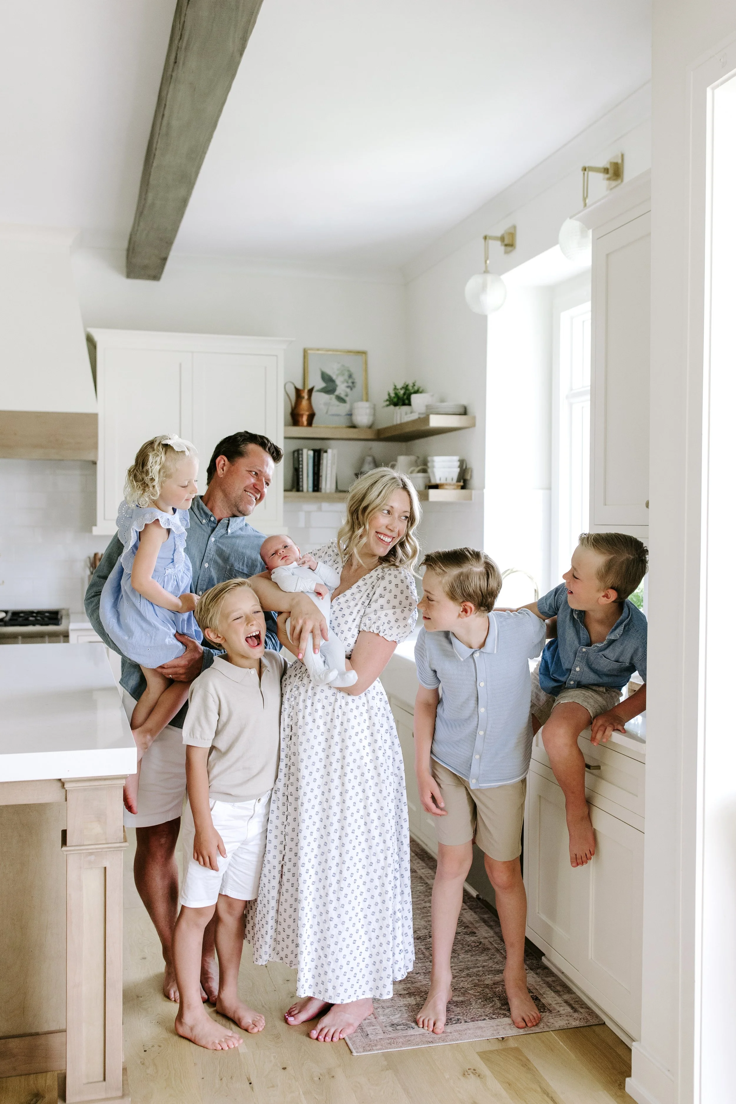 A family gathered in a bright kitchen, smiling and holding their children, with a mix of adults and kids, all smiling and enjoying each other's company.
