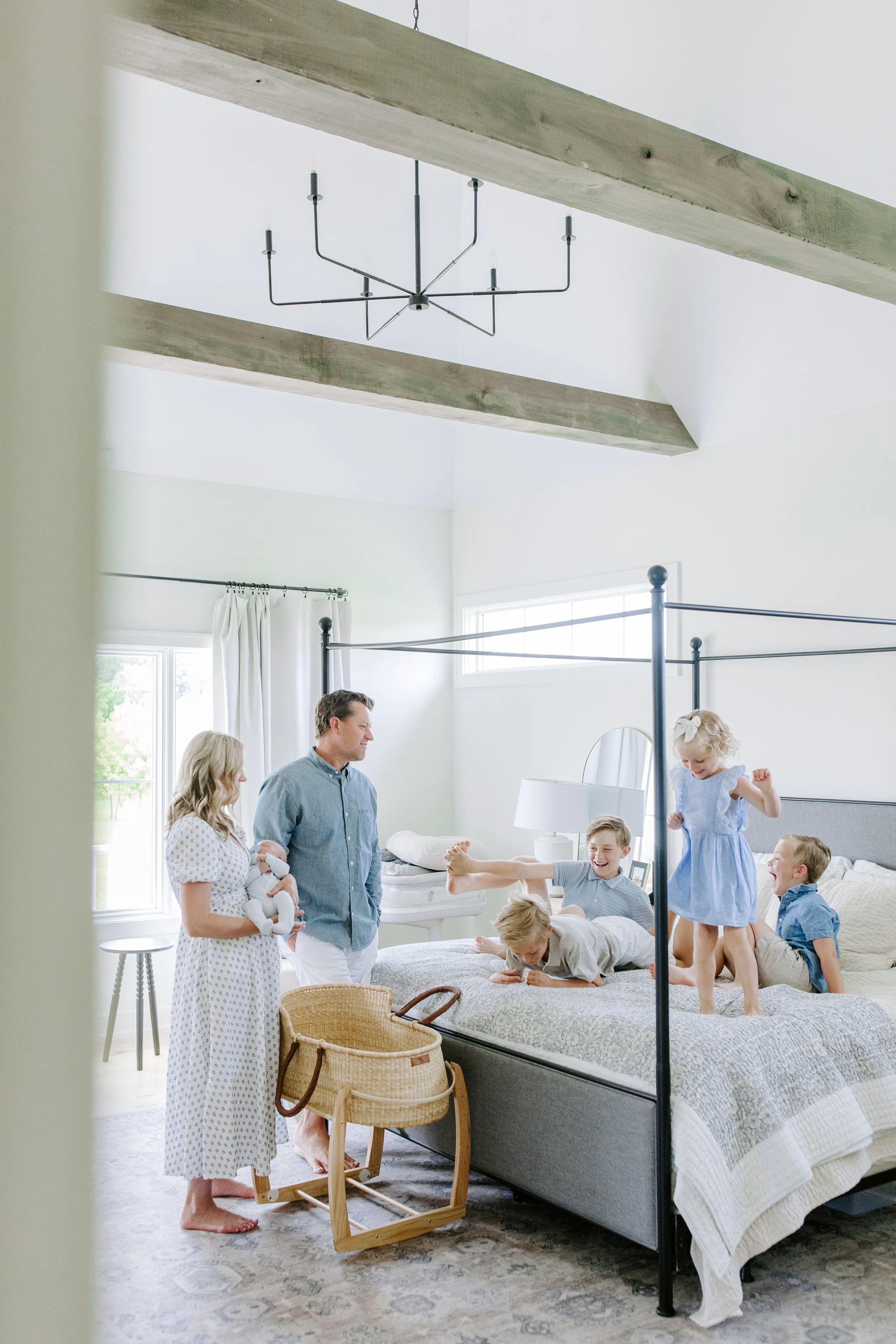 A family gathers in a bright, white-bedroom with a four-poster bed where children are playing and jumping. Adults are observing, with one woman holding a baby. A ceiling light fixture is visible. The room has large windows with white curtains, and a 