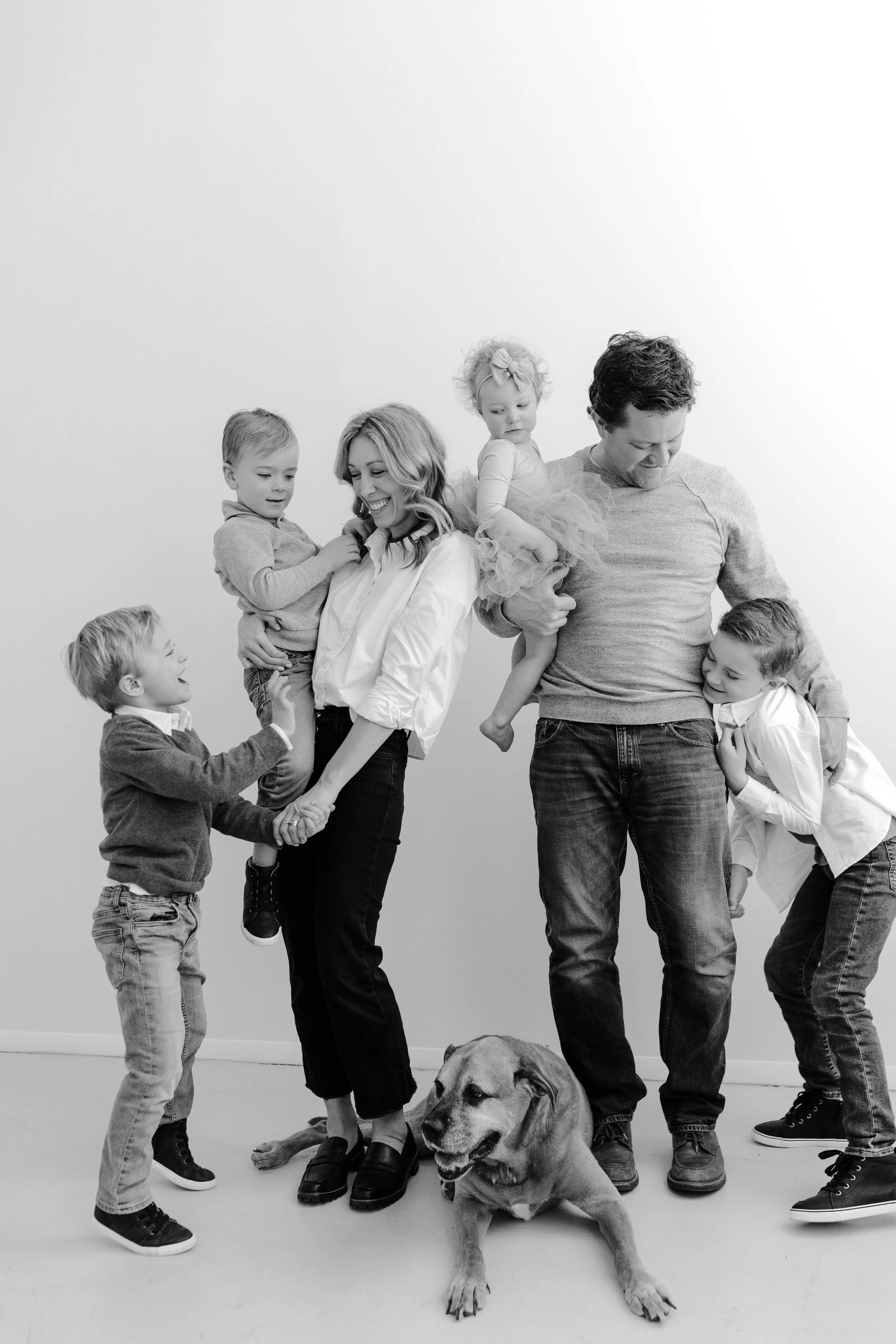 A family of four children and two adults, possibly parents, smiling and playing with each other, with a dog lying on the floor, in front of a plain white background.
