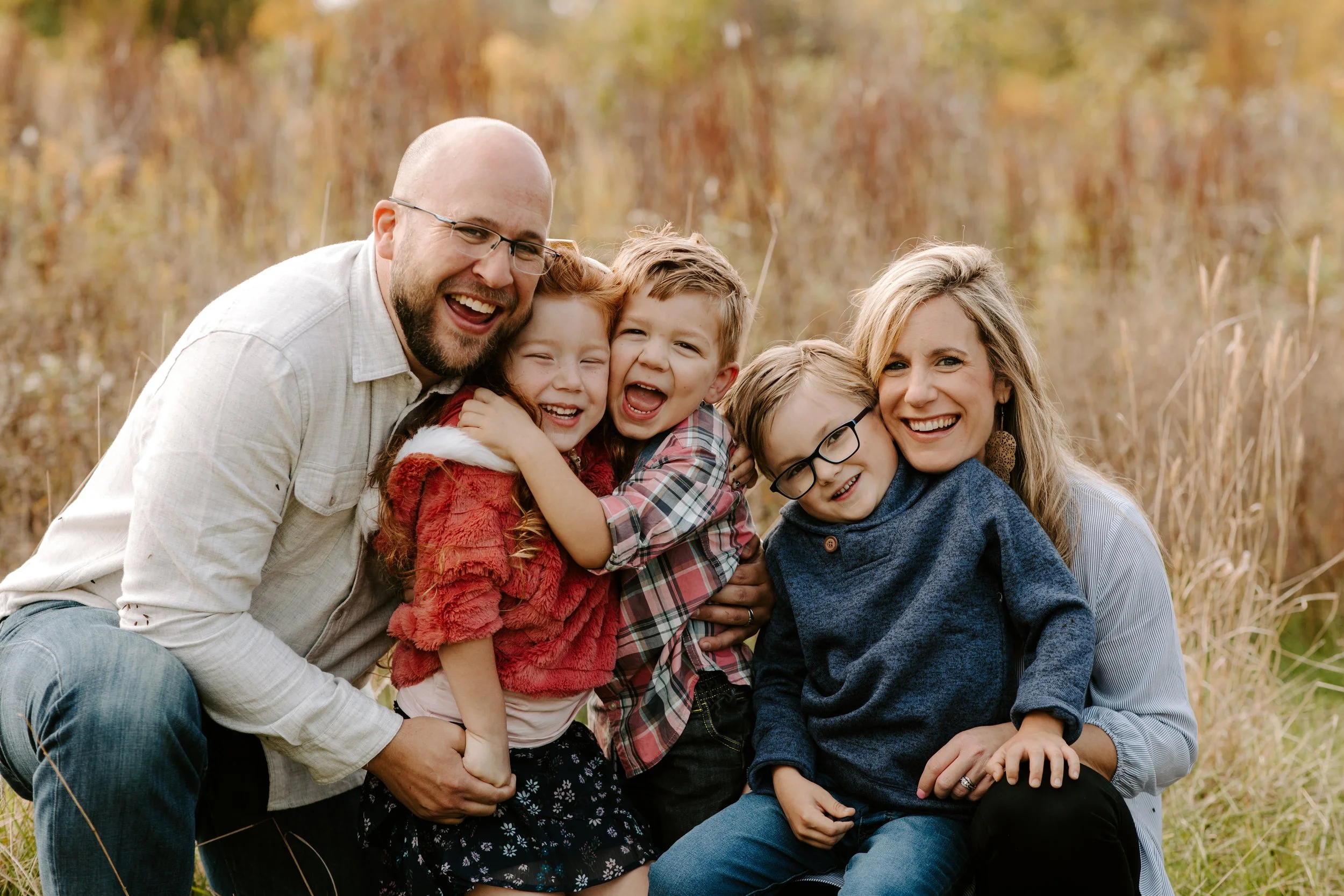A happy family of six with two adults and four children, smiling and embracing outdoors in a field with fall foliage in the background.