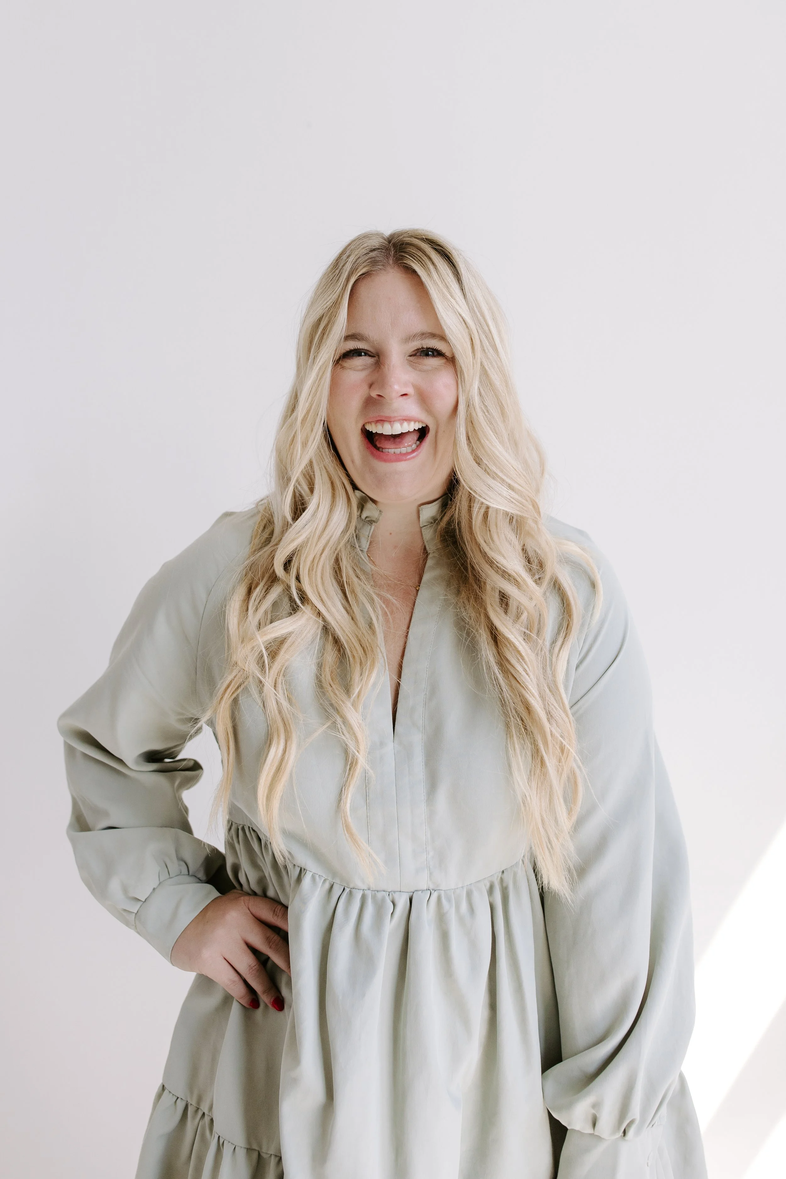 A woman with long blonde hair, smiling and laughing, wearing a light-colored dress with long sleeves, standing against a plain white background.