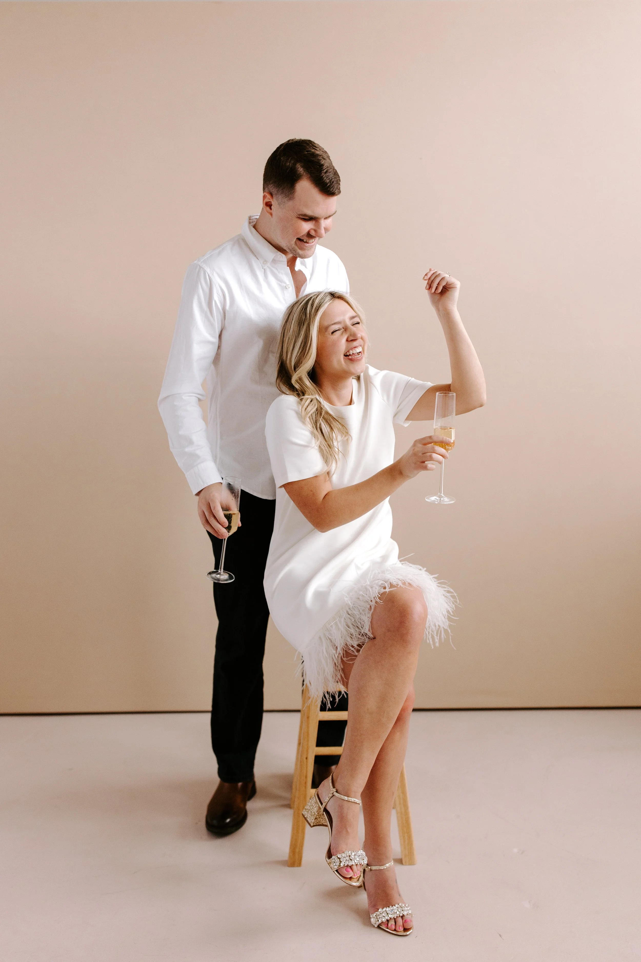 A man and woman celebrate with champagne, the woman is sitting on a stool and the man stands behind her, both smiling and laughing.