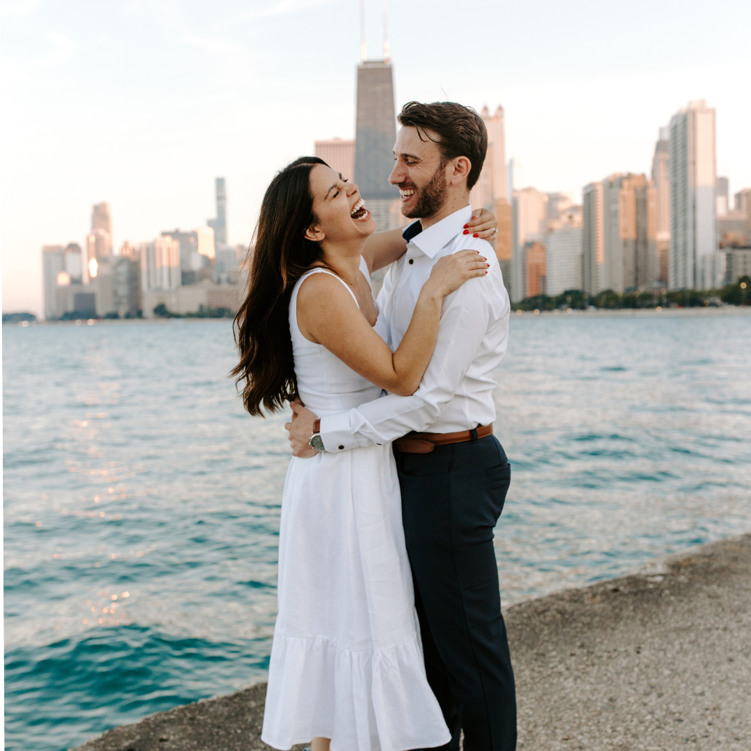 A couple in white and dark clothing embracing and smiling by the water with a city skyline in the background.