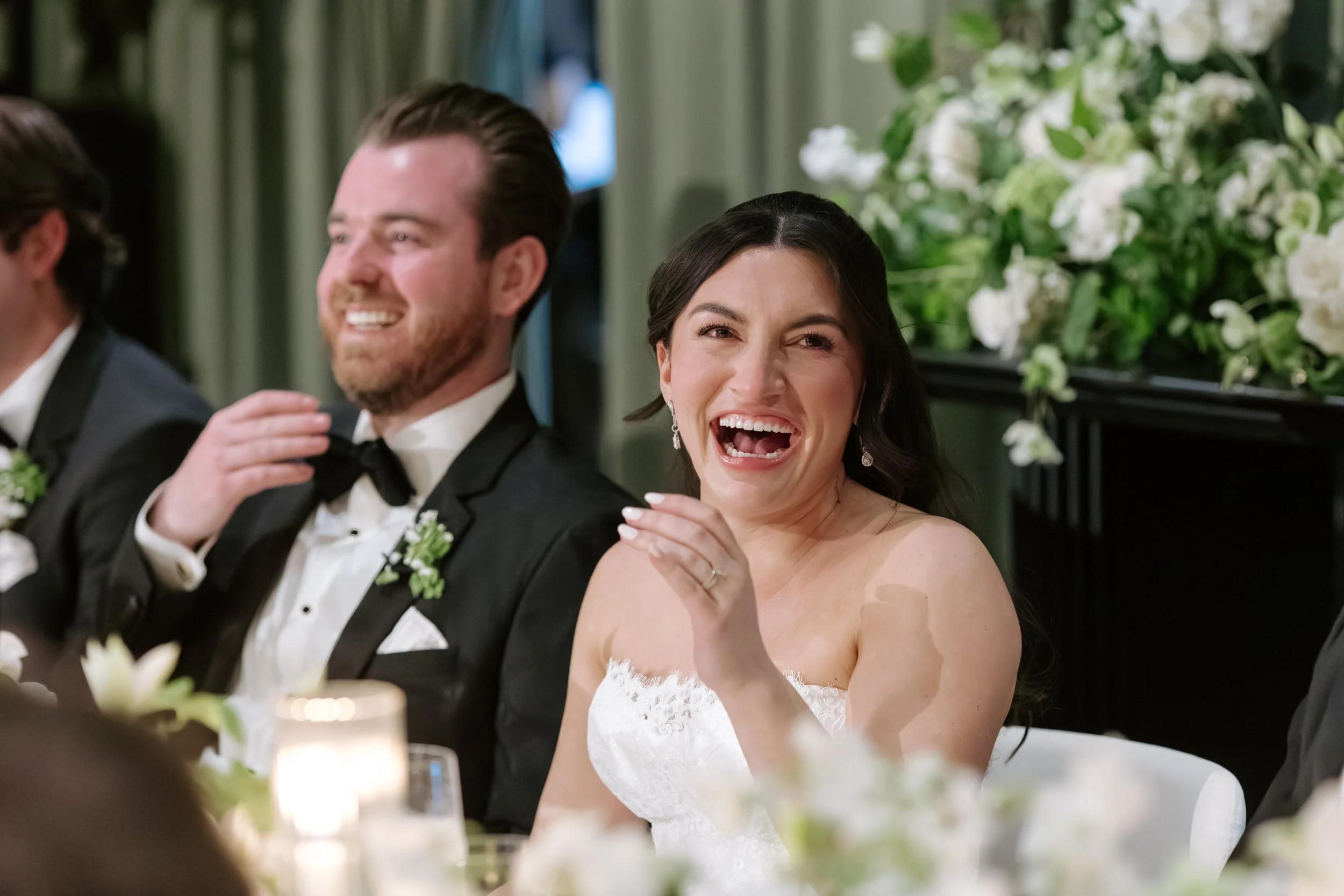 A bride and groom sitting at a wedding reception, smiling and enjoying the moment, with floral decorations in the background.