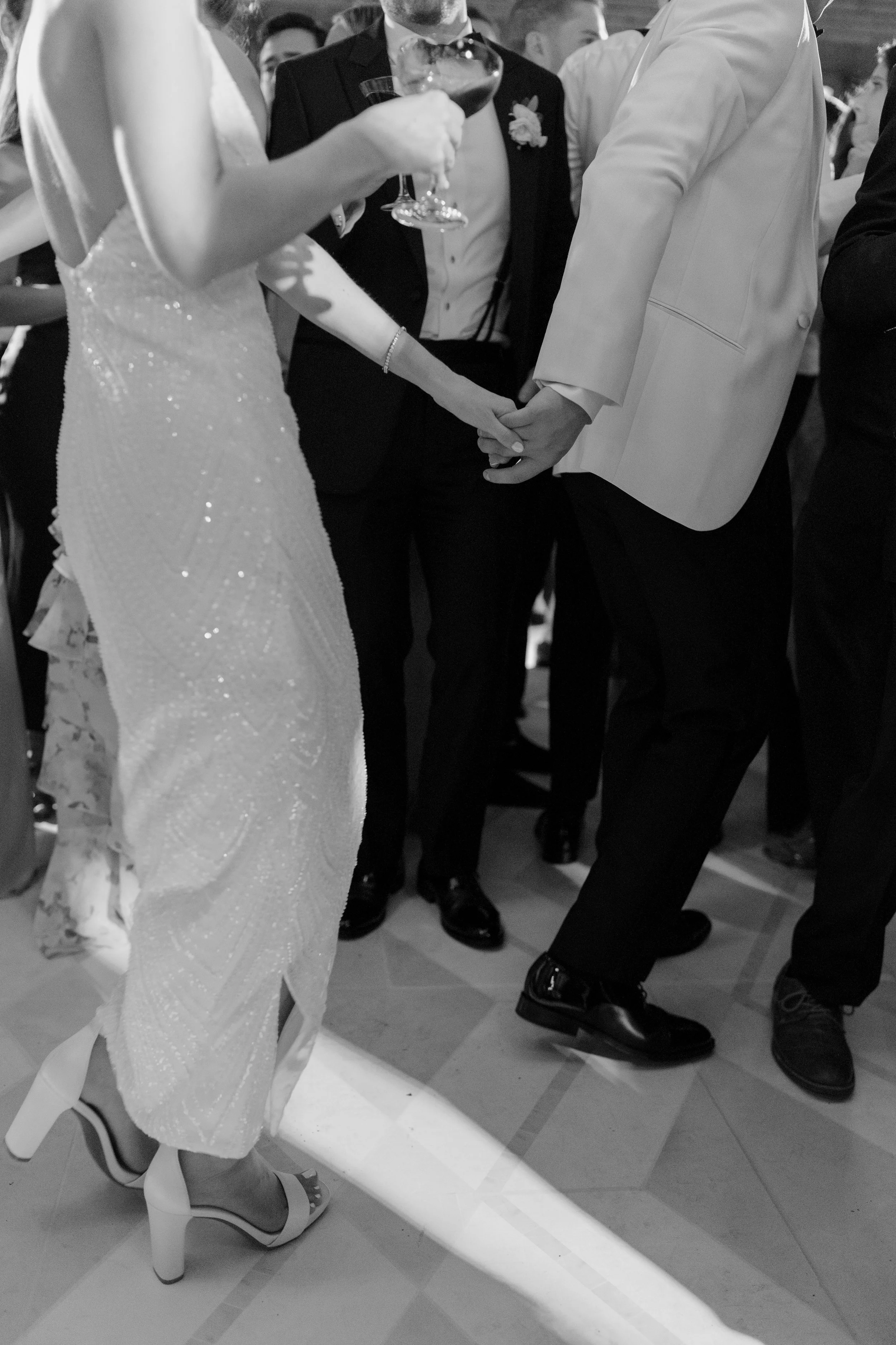 A black and white photo of a wedding reception, showing the bride and groom holding hands on the dance floor, surrounded by guests. The bride wears a sparkling wedding dress and high heels, and the groom wears a white tuxedo jacket and dark shoes. On