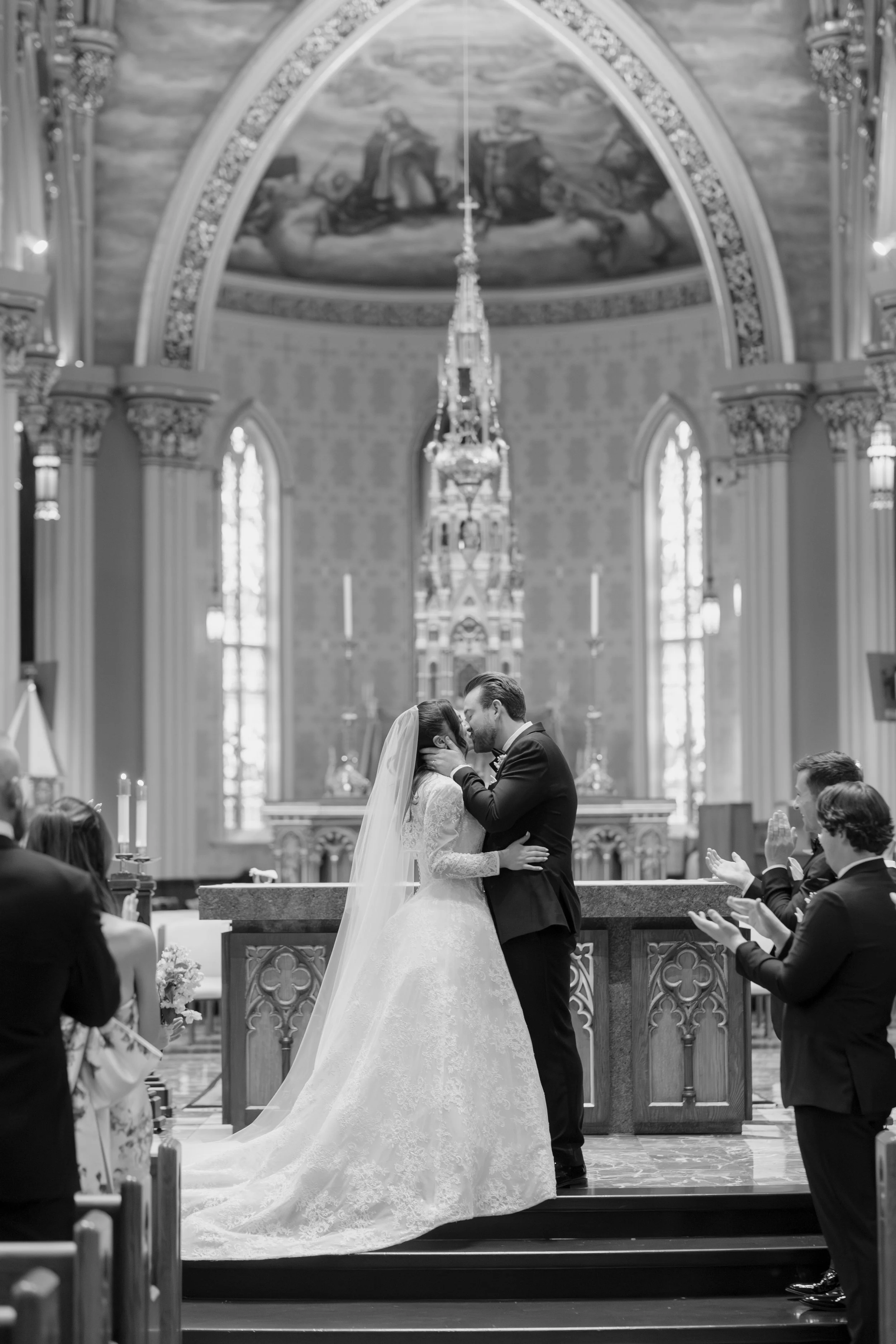 A bride and groom kiss at their wedding ceremony inside a church with high, arched ceilings and stained glass windows, surrounded by wedding guests clapping and celebrating.