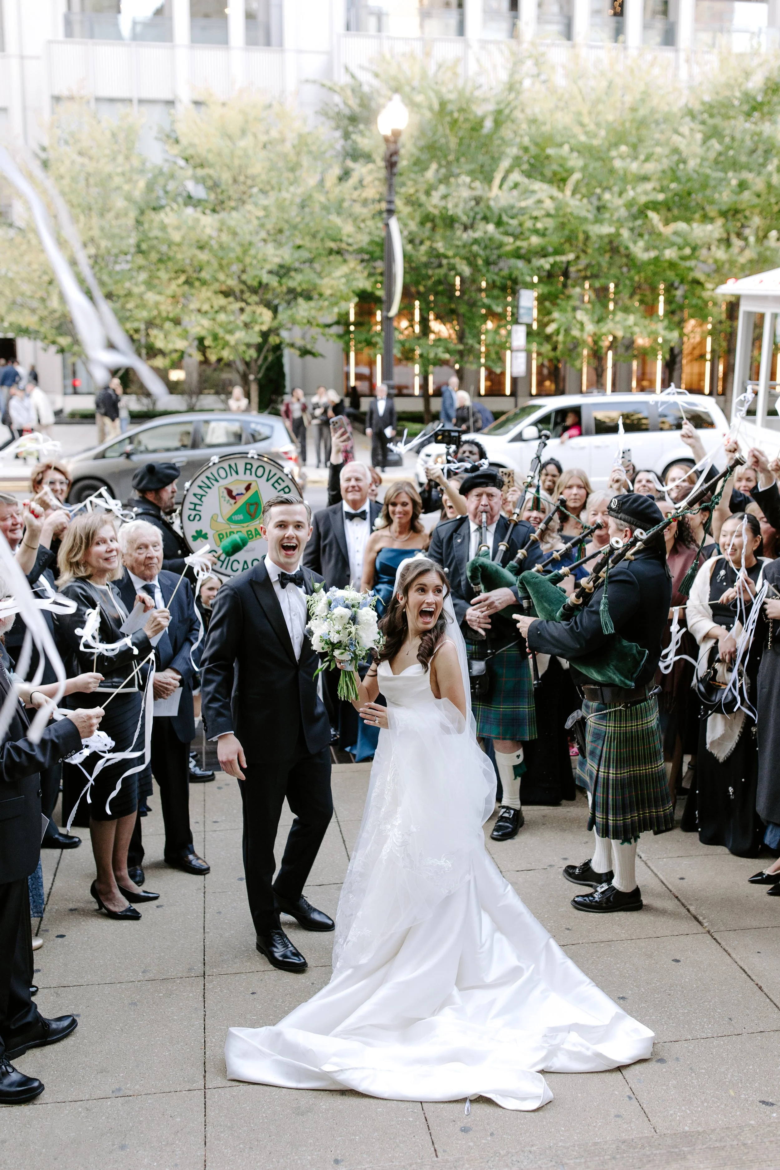 A newly married couple in wedding attire walking down a city sidewalk surrounded by guests celebrating with ribbon and streamers. The groom is smiling and holding the bride's hand as she holds a bouquet of white flowers. Guests are gathered around, s
