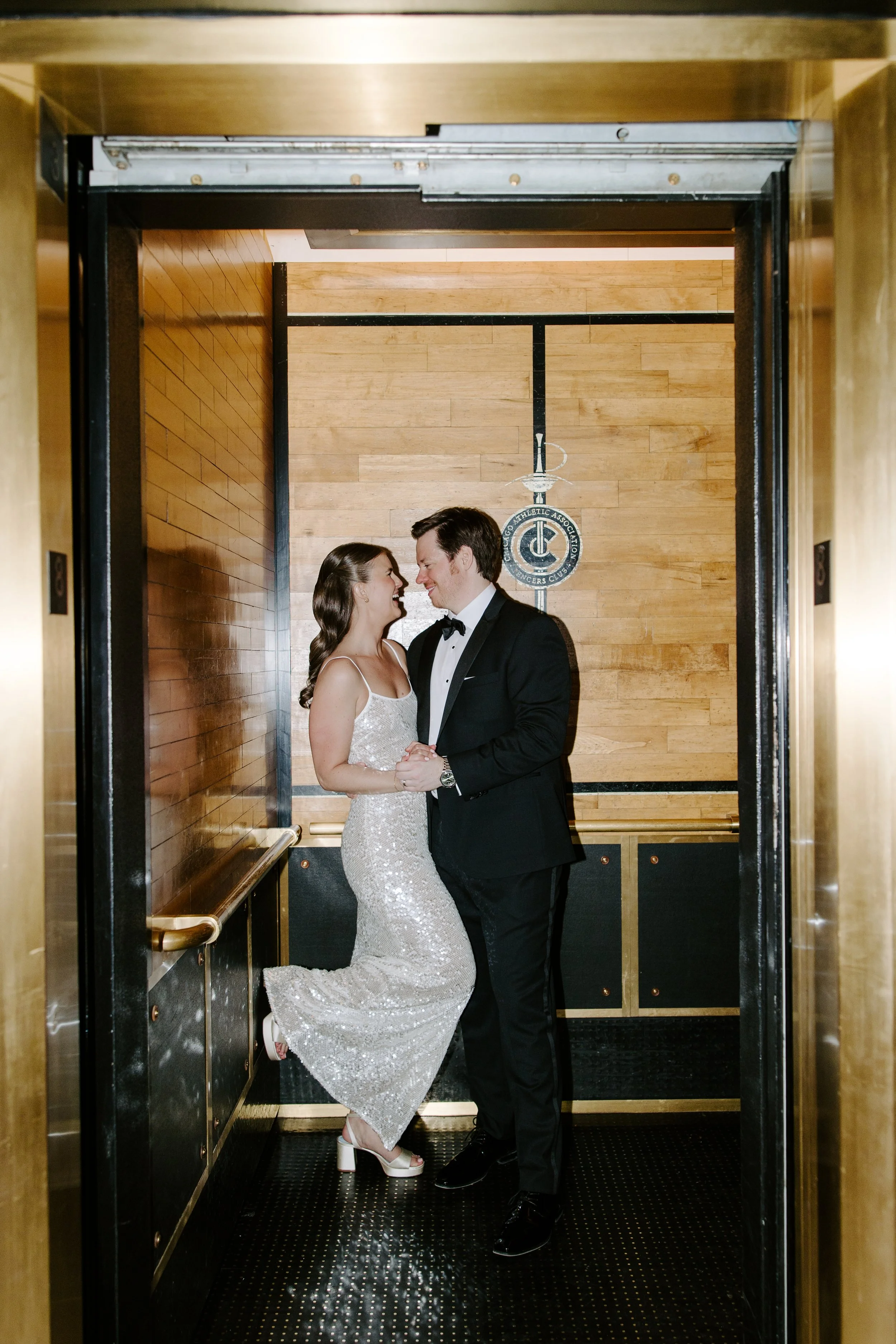 A couple dressed in formal attire, the woman in a sparkling white gown and the man in a tuxedo, standing inside an elevator with wooden paneling on the walls.