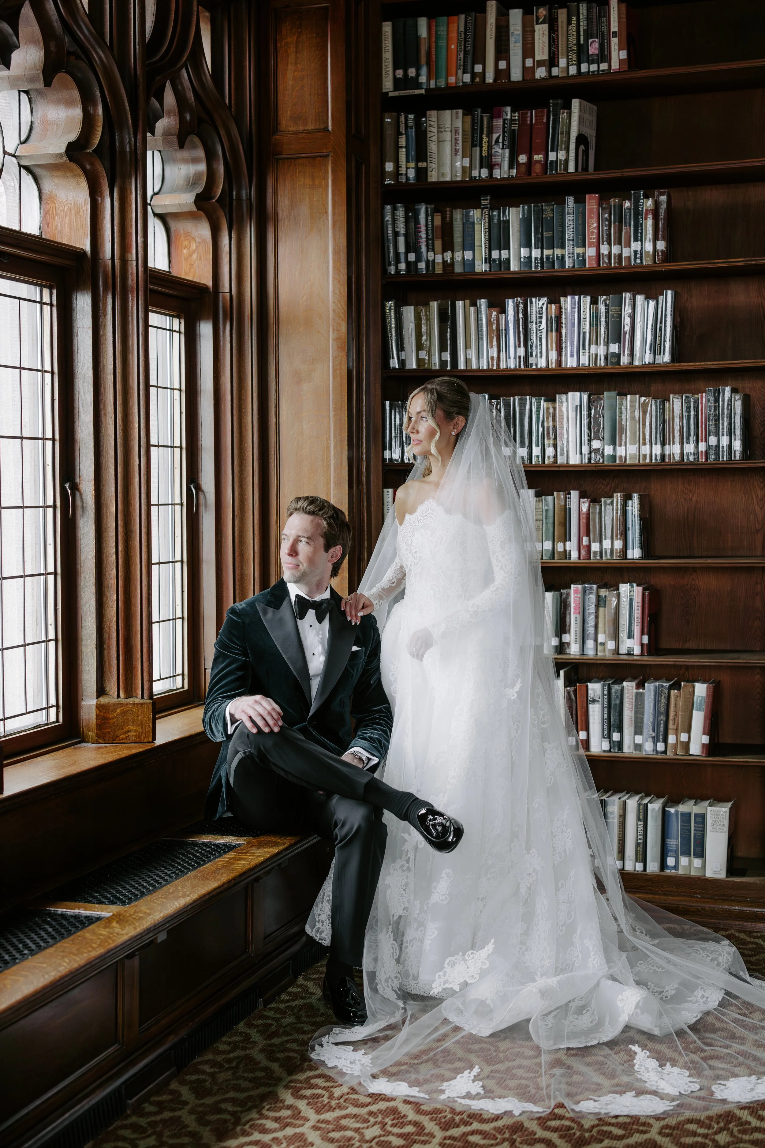 A bride and groom in wedding attire in a historic library with tall wooden bookshelves and large stained glass windows. The groom sits on a bench by the window, looking up, while the bride stands beside him, wearing a lace wedding gown and veil.