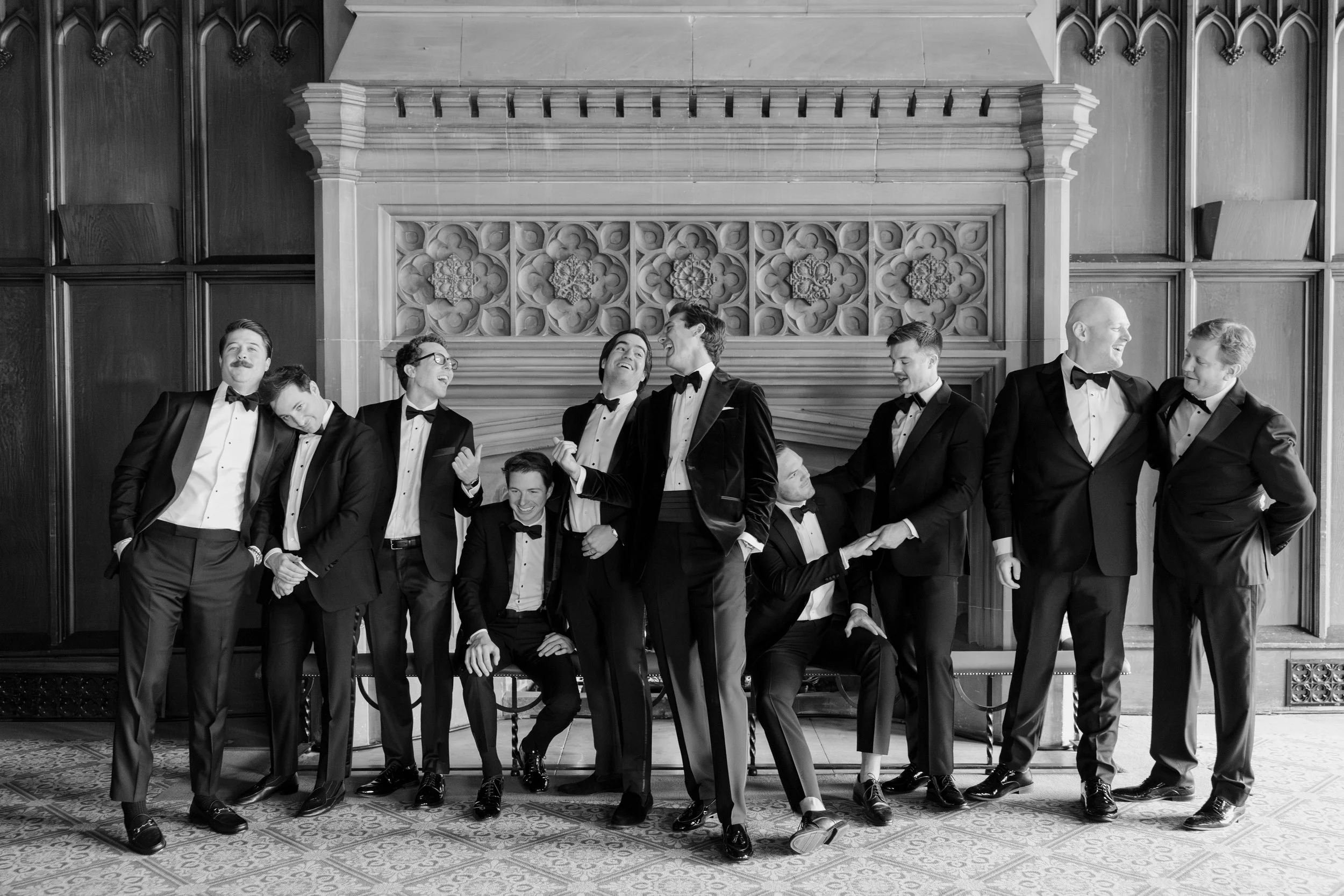 Group of men in tuxedos smiling and interacting in a formal setting with ornate wood paneling and decorative architectural features