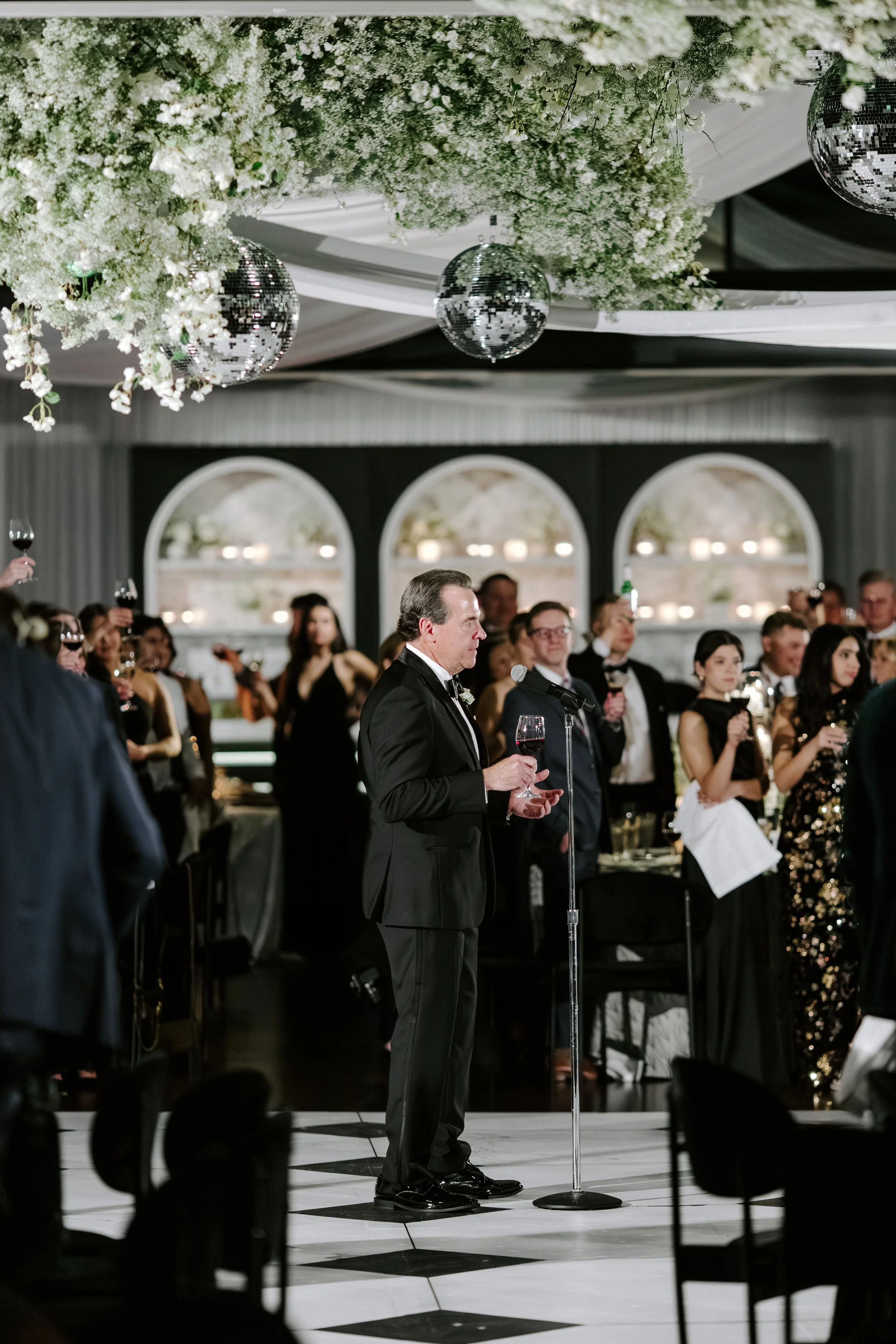 A man in a tuxedo standing with a glass of red wine at a formal event while others host and guest stand behind a microphone in a decorated banquet hall with floral arrangements and disco balls hanging from the ceiling.