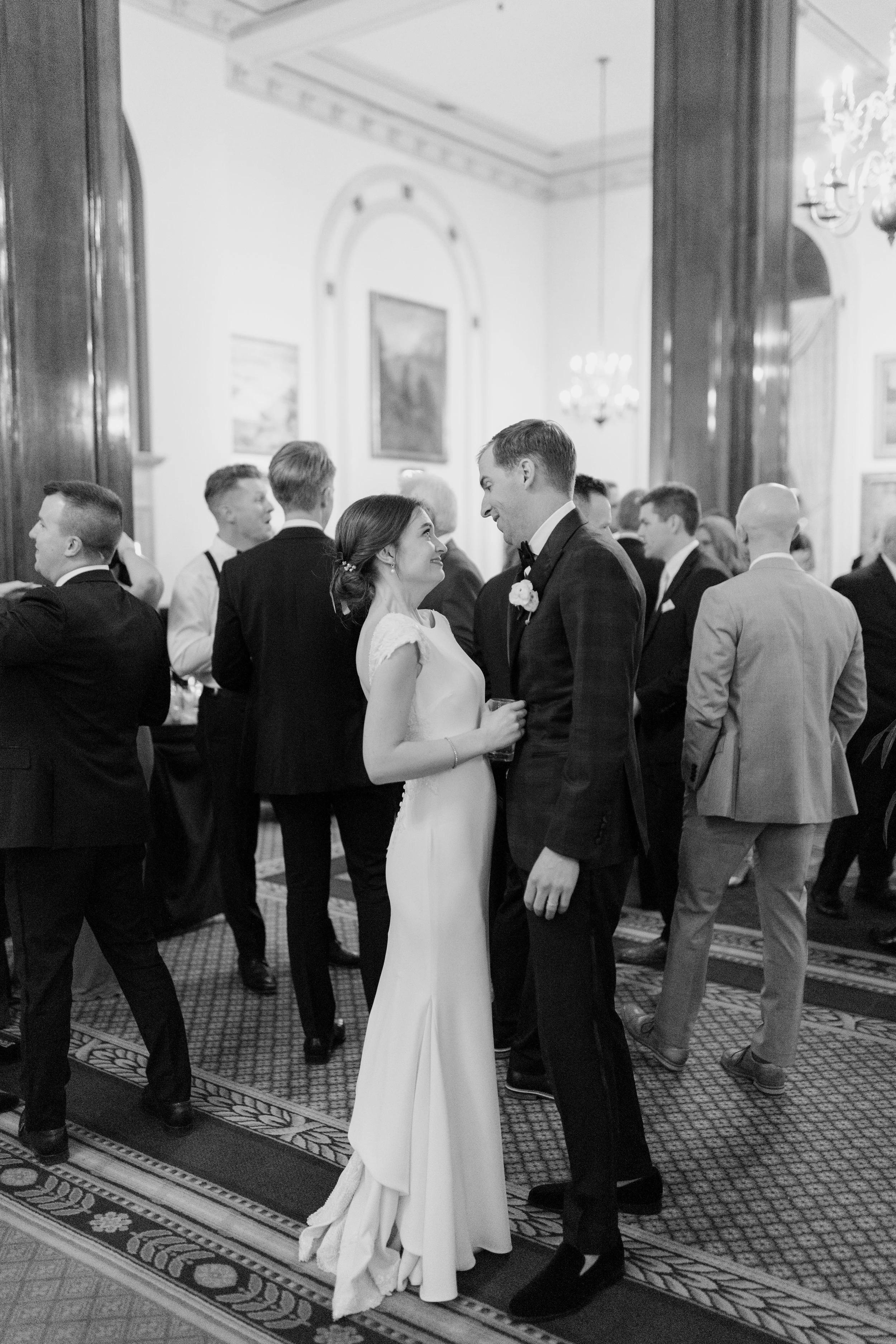 A bride and groom are dancing and looking into each other's eyes at a wedding reception in an elegant, decorated hall with chandeliers and framed artwork on the walls. The bride is wearing a white gown and the groom is wearing a dark suit with a bow 