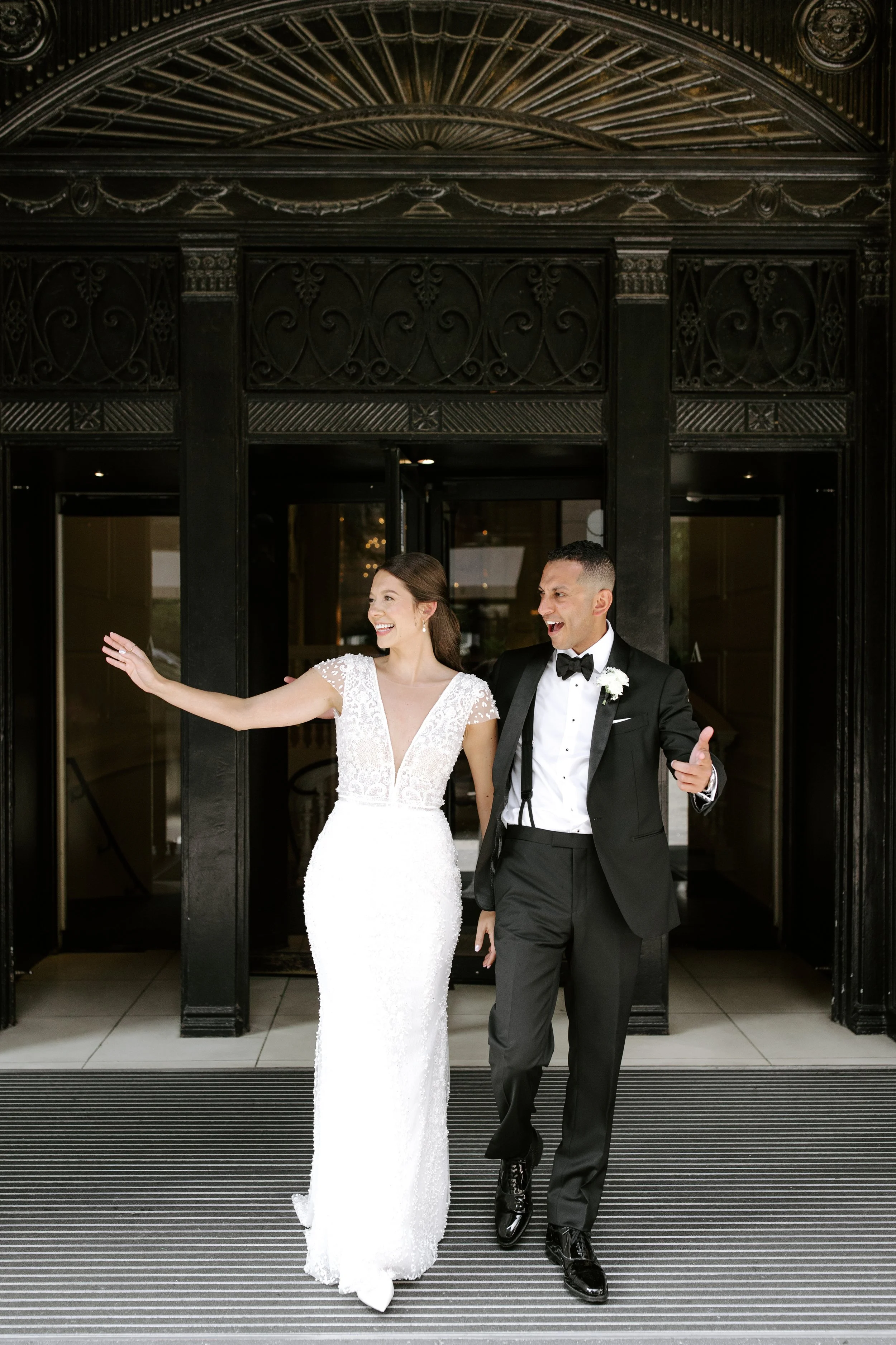 A bride and groom walking out of a building together, smiling and happily celebrating their wedding day. The bride is wearing a white lace wedding dress, and the groom is dressed in a black tuxedo with a bow tie.