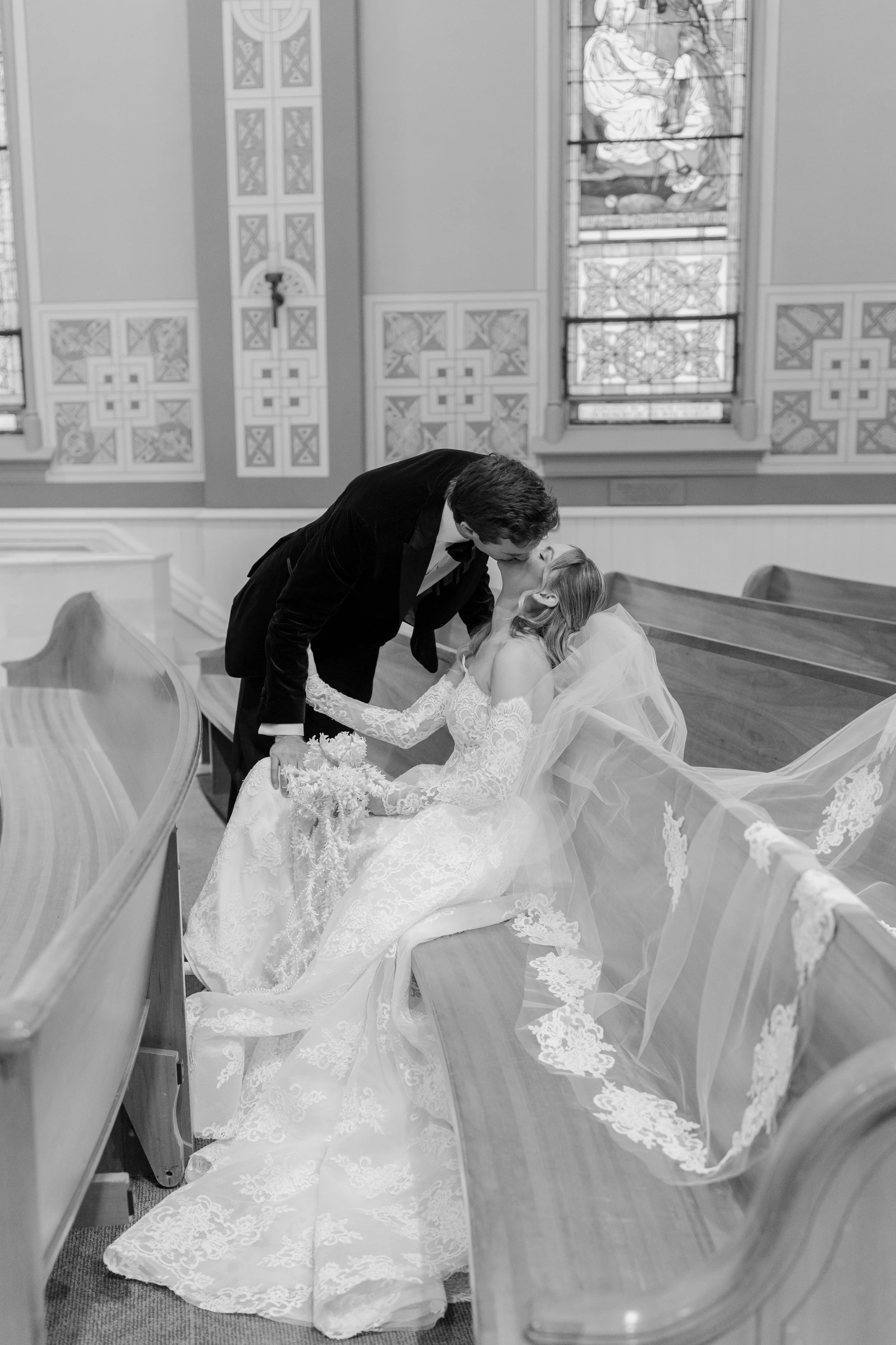 A man and a woman in a church, with the woman in a wedding dress and veil sitting on a pew, and the man leaning over, kissing her, holding her hand, with stained glass windows in the background.