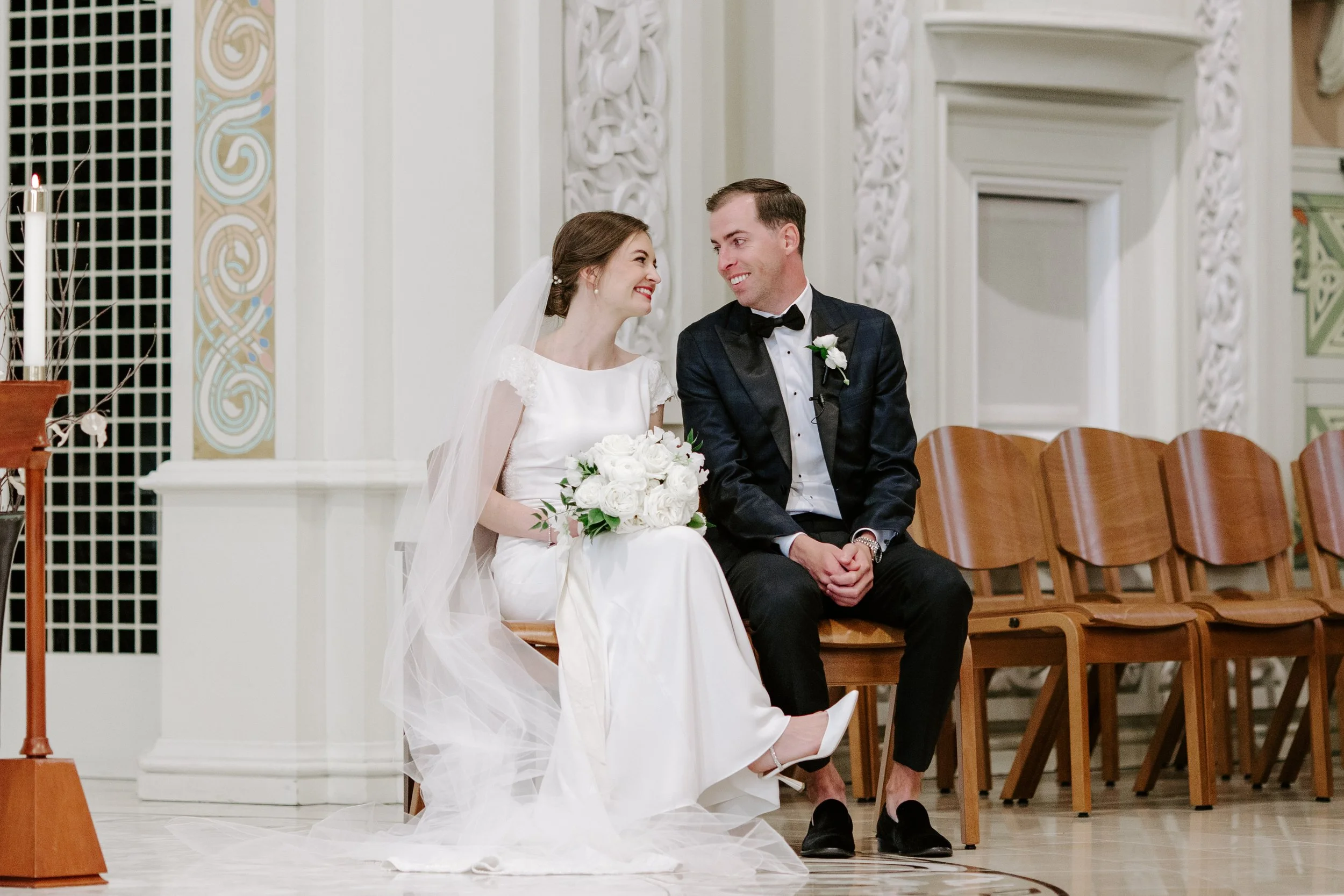 A bride and groom sit together during a wedding ceremony in a church, smiling and looking at each other. The bride is wearing a white wedding dress and veil, holding a bouquet of white flowers. The groom is in a dark tuxedo with a bow tie and a white