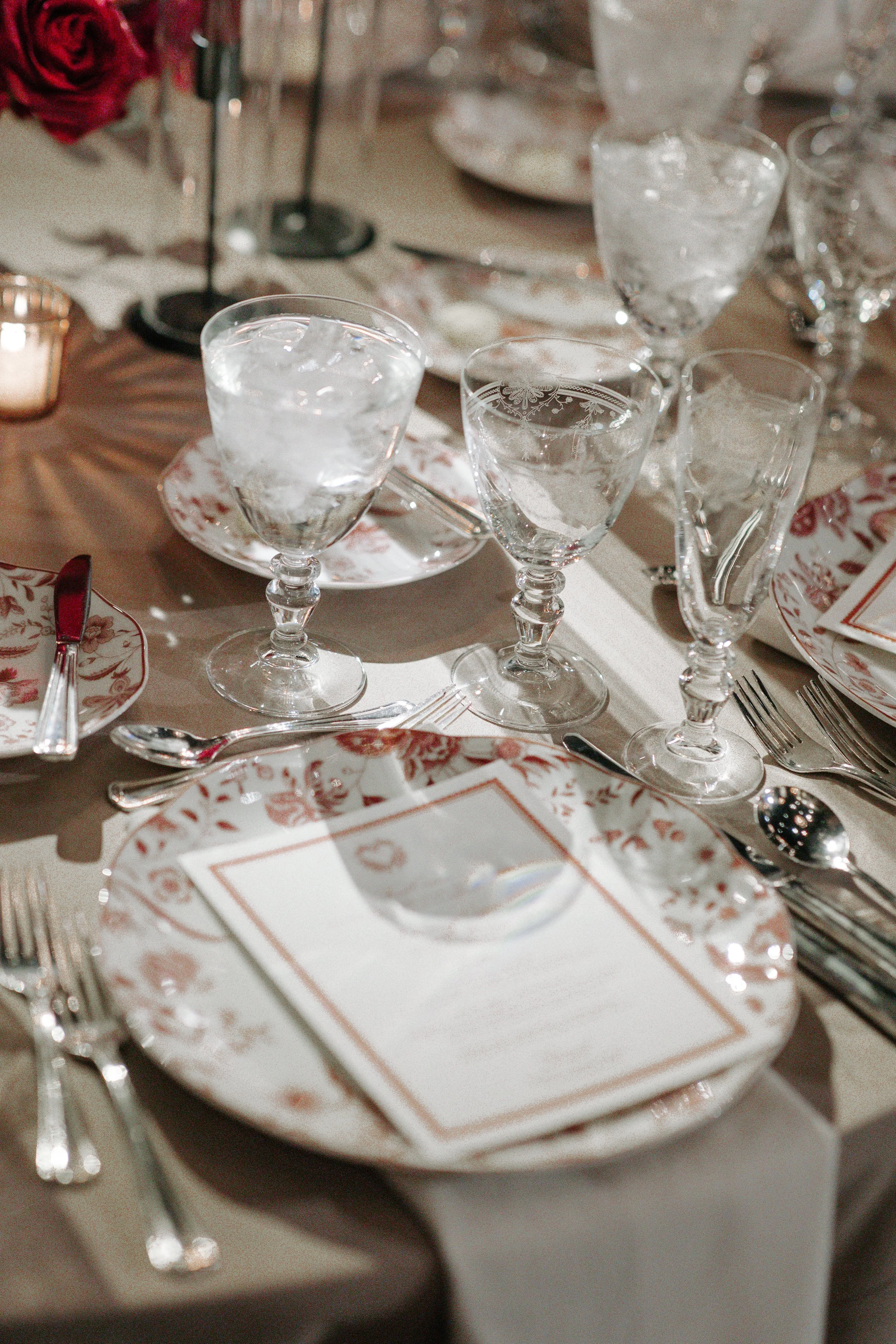 Elegant table setting with glassware, plates, silverware, and a menu or place card on a tablecloth, likely at a formal event or dinner party.
