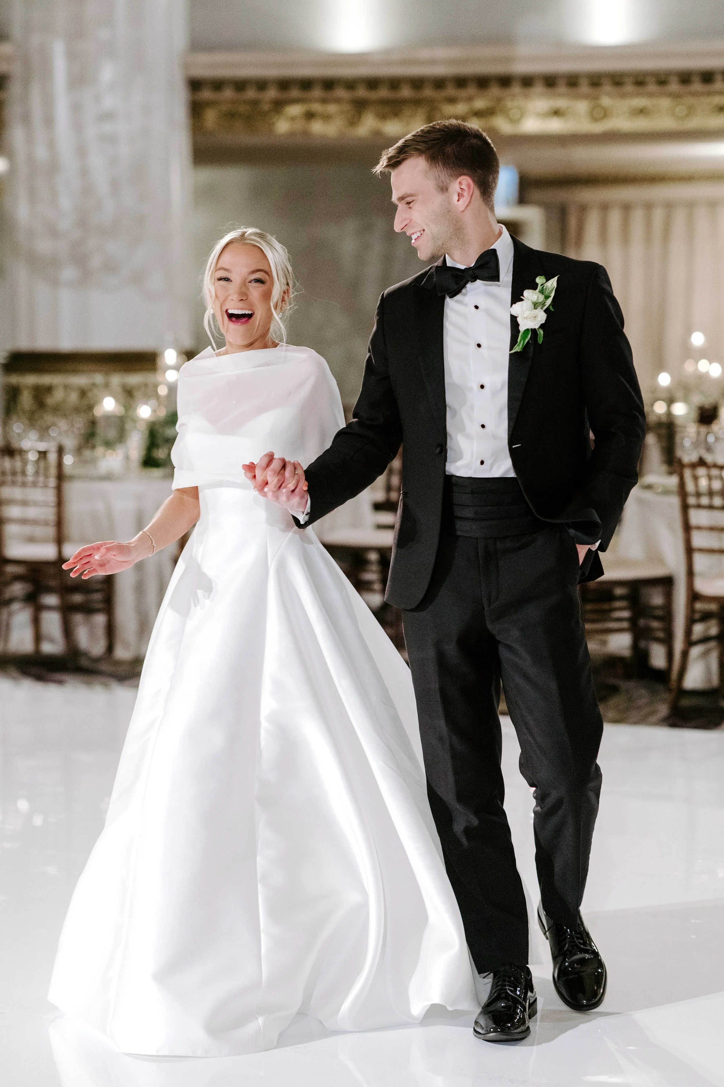 A bride and groom dancing at their wedding reception in an elegant ballroom, smiling and holding hands.