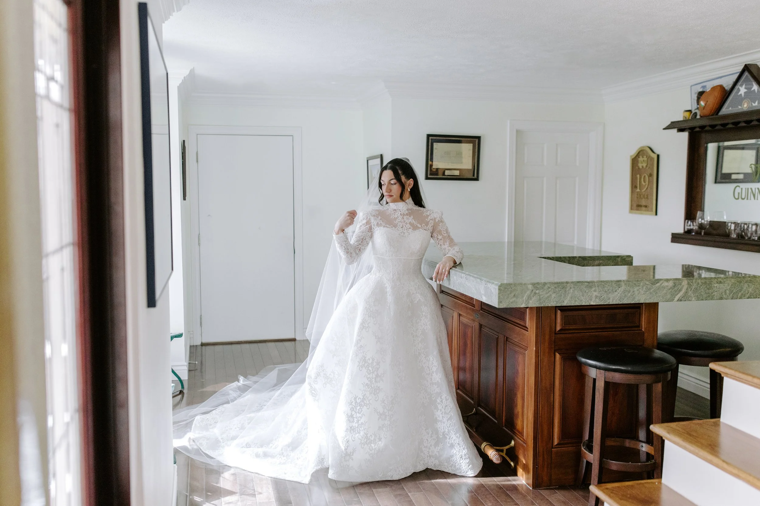 A woman in a white lace wedding dress with a train standing in a kitchen, looking down with her head turned slightly to the side, near a marble-topped counter.