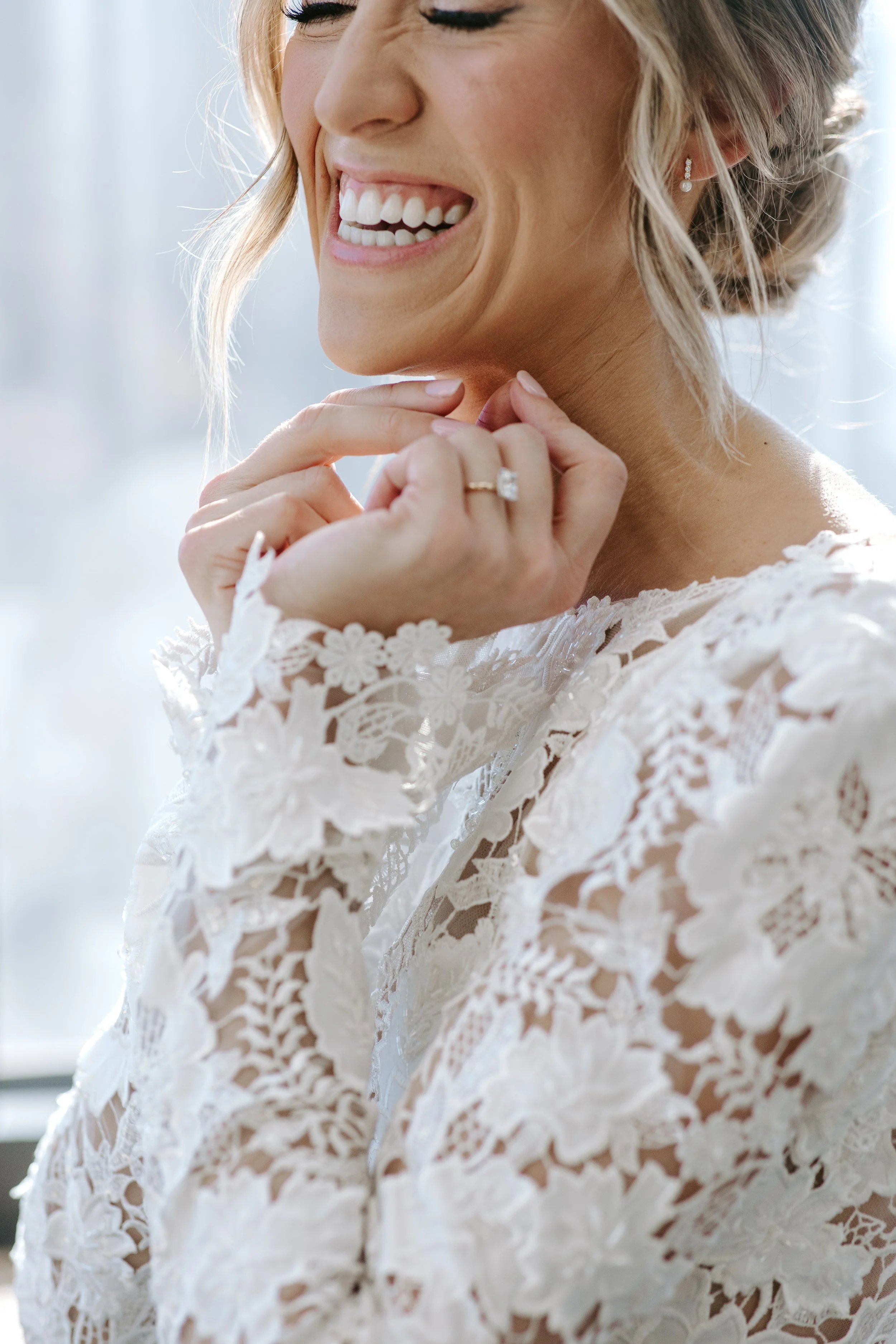 Close-up of a woman with a smile, wearing pearl earrings and a ring, dressed in lace with a floral pattern, showcasing her face and hands.