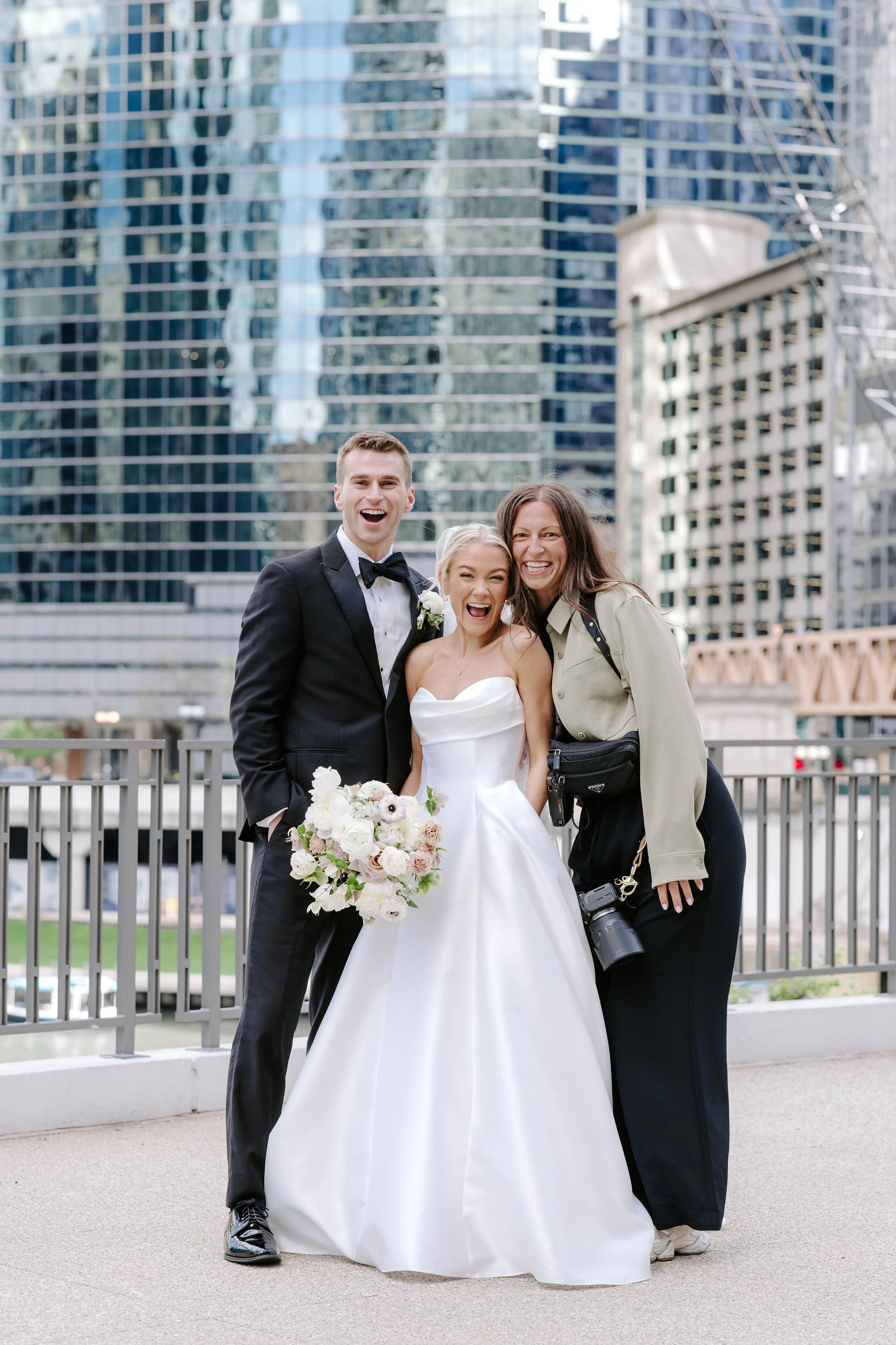 A bride in a white wedding dress holding a bouquet, with a man in a tuxedo and a woman in casual clothing standing beside her, all smiling happily outside with modern glass buildings in the background.