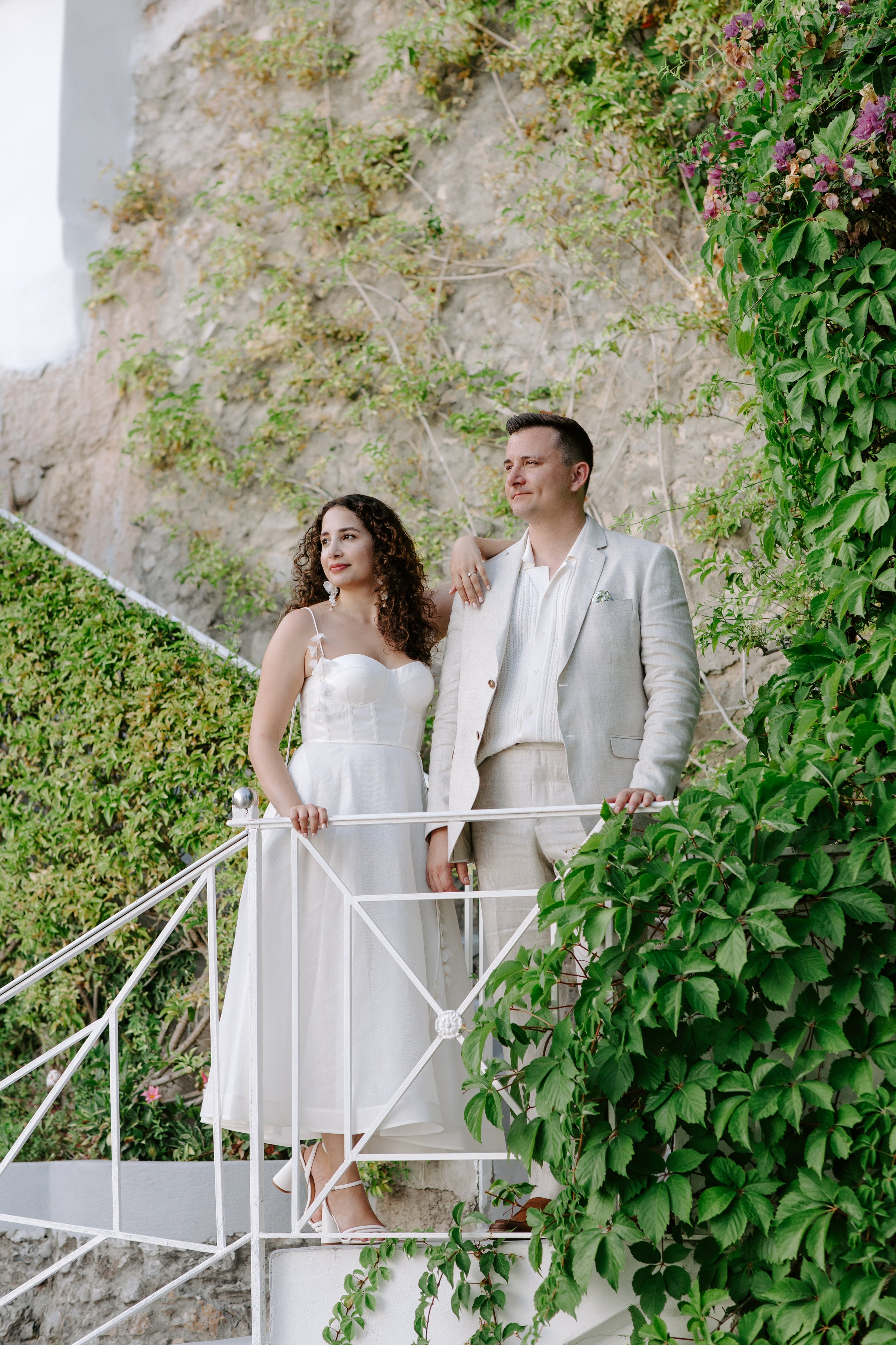 A man and woman dressed in wedding attire standing on a white balcony with greenery and a stone wall in the background.