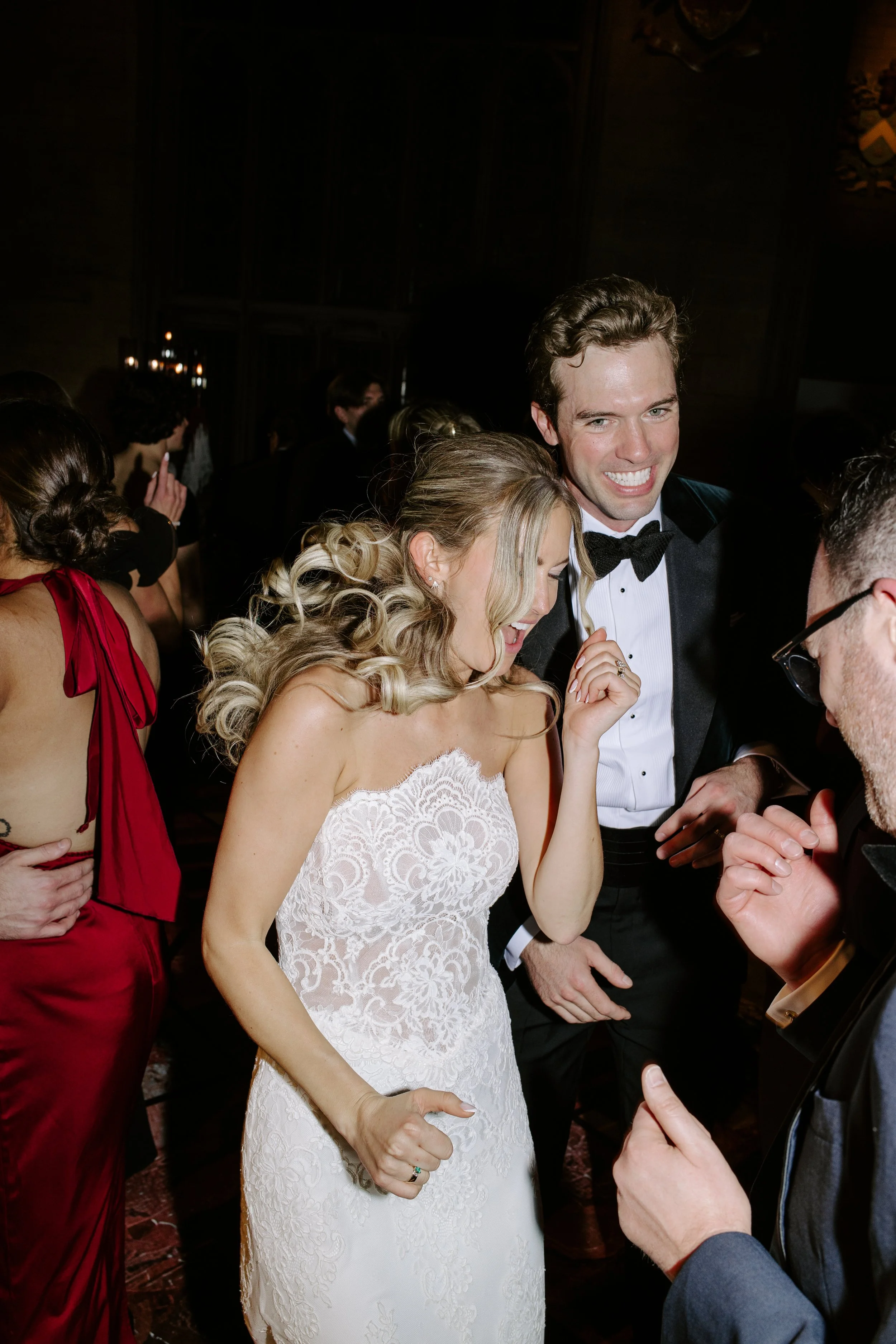 A bride in a white lace wedding dress smiling and laughing with a groom wearing a tuxedo at a wedding reception. They are surrounded by guests, some in formal attire, in a dimly lit indoor setting.