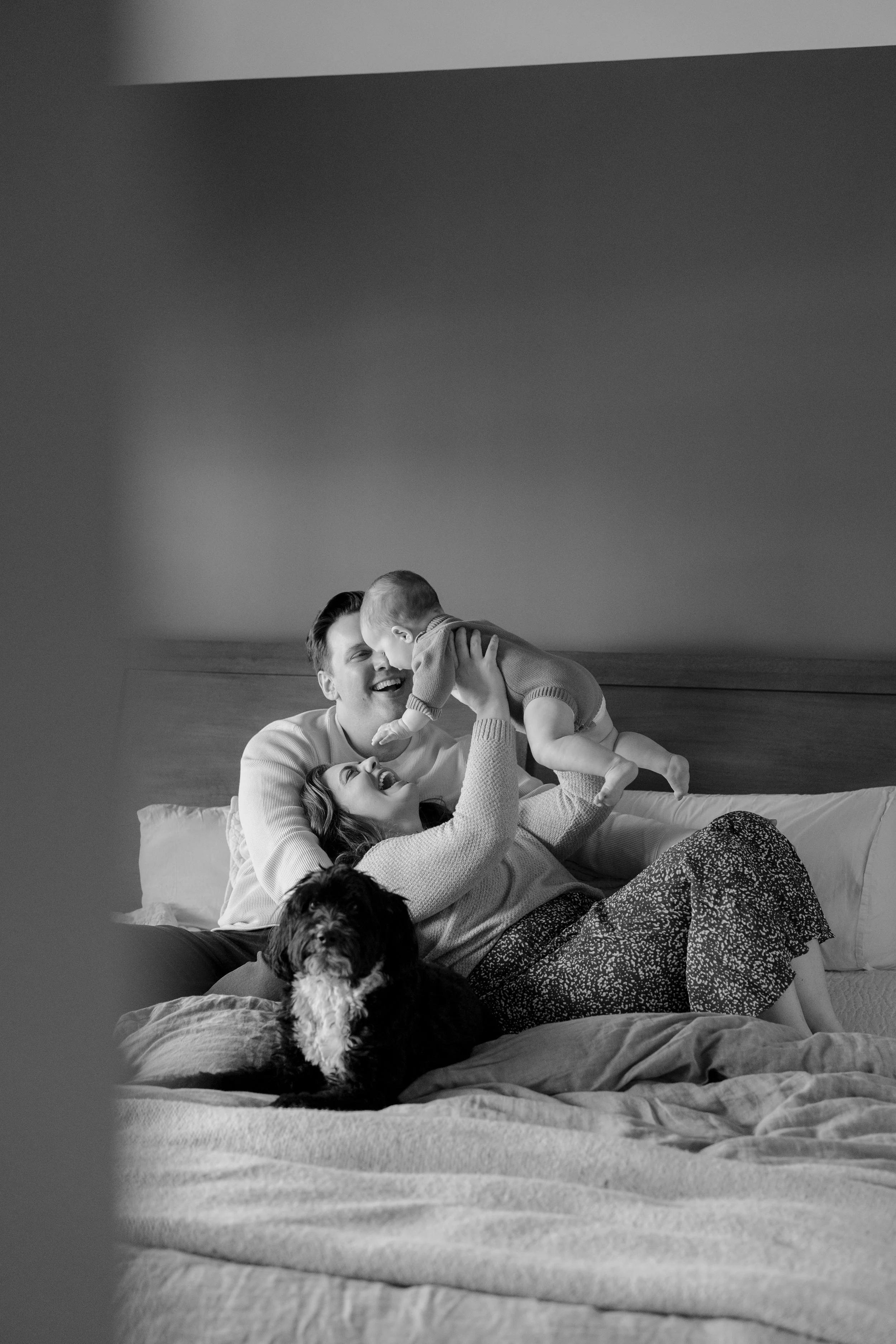 A happy family of three people and an adorable dog playing together on a bed in a cozy room, captured in black and white.