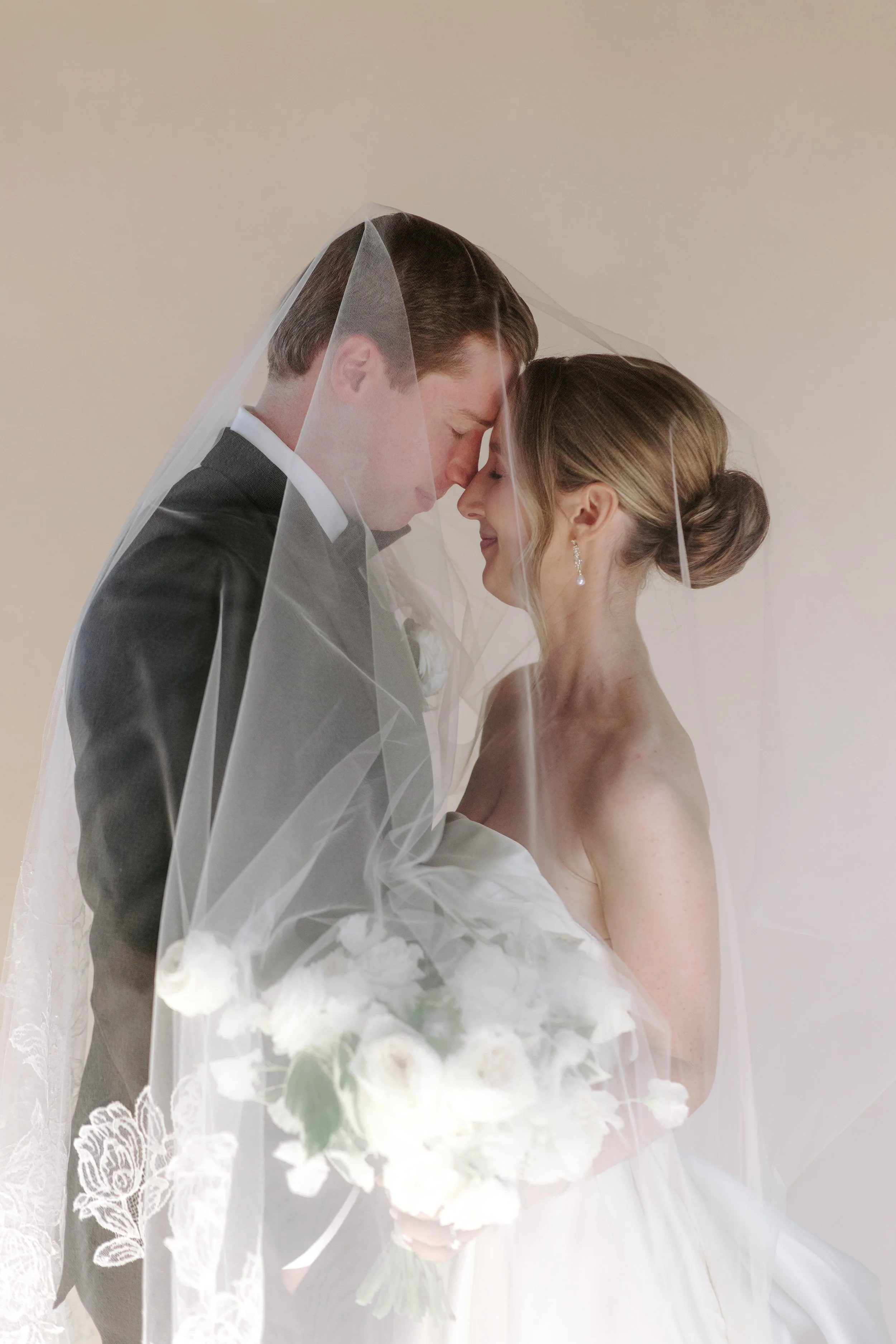 A bride and groom under a sheer veil, close together with foreheads touching, holding a bouquet of white flowers, against a plain background.