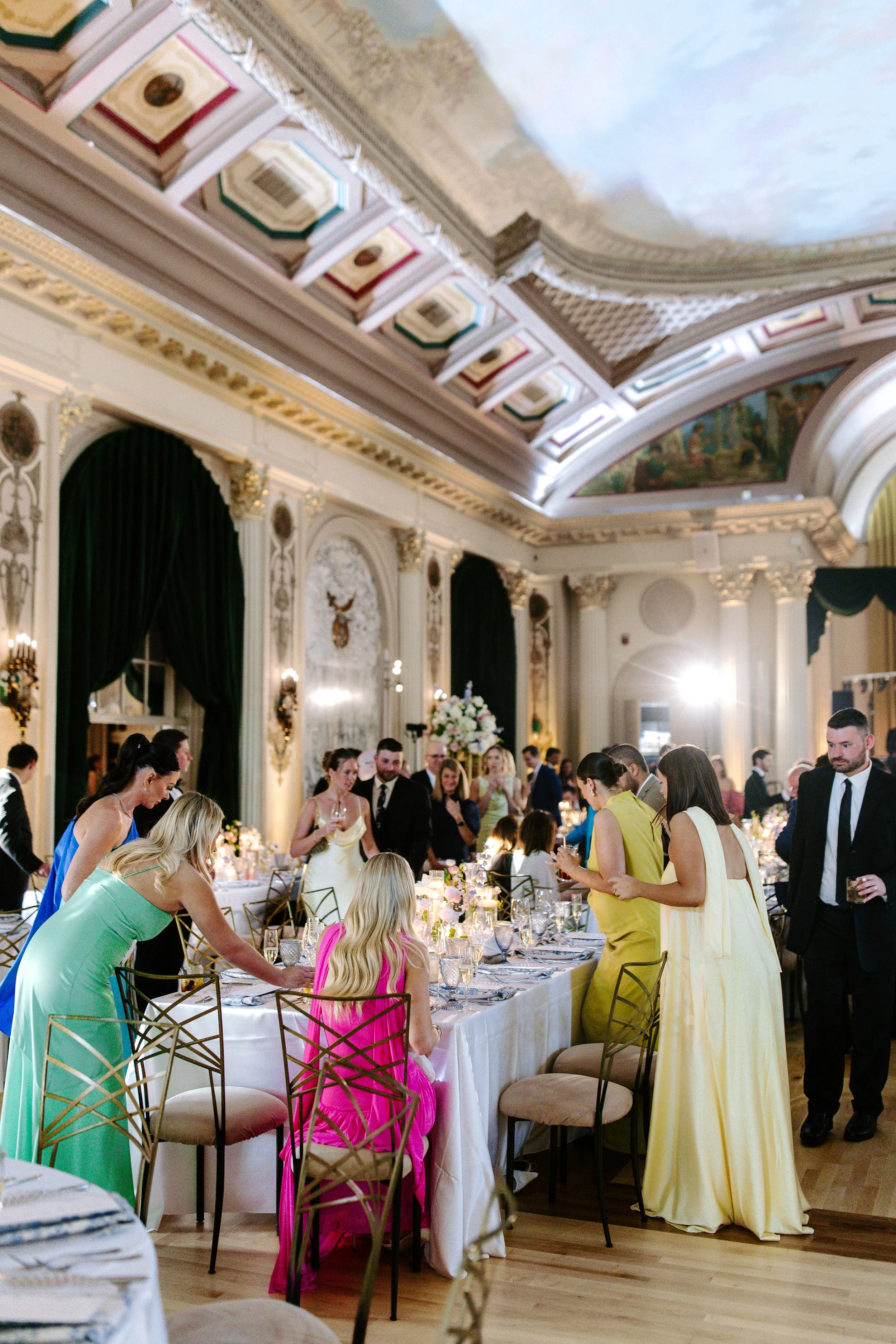 A formal gathering or banquet in an elegant, ornate room with high ceilings, large curtains, and detailed architectural decor. Guests are dressed in formal attire, standing around a long table set with silverware, candles, and floral centerpieces.
