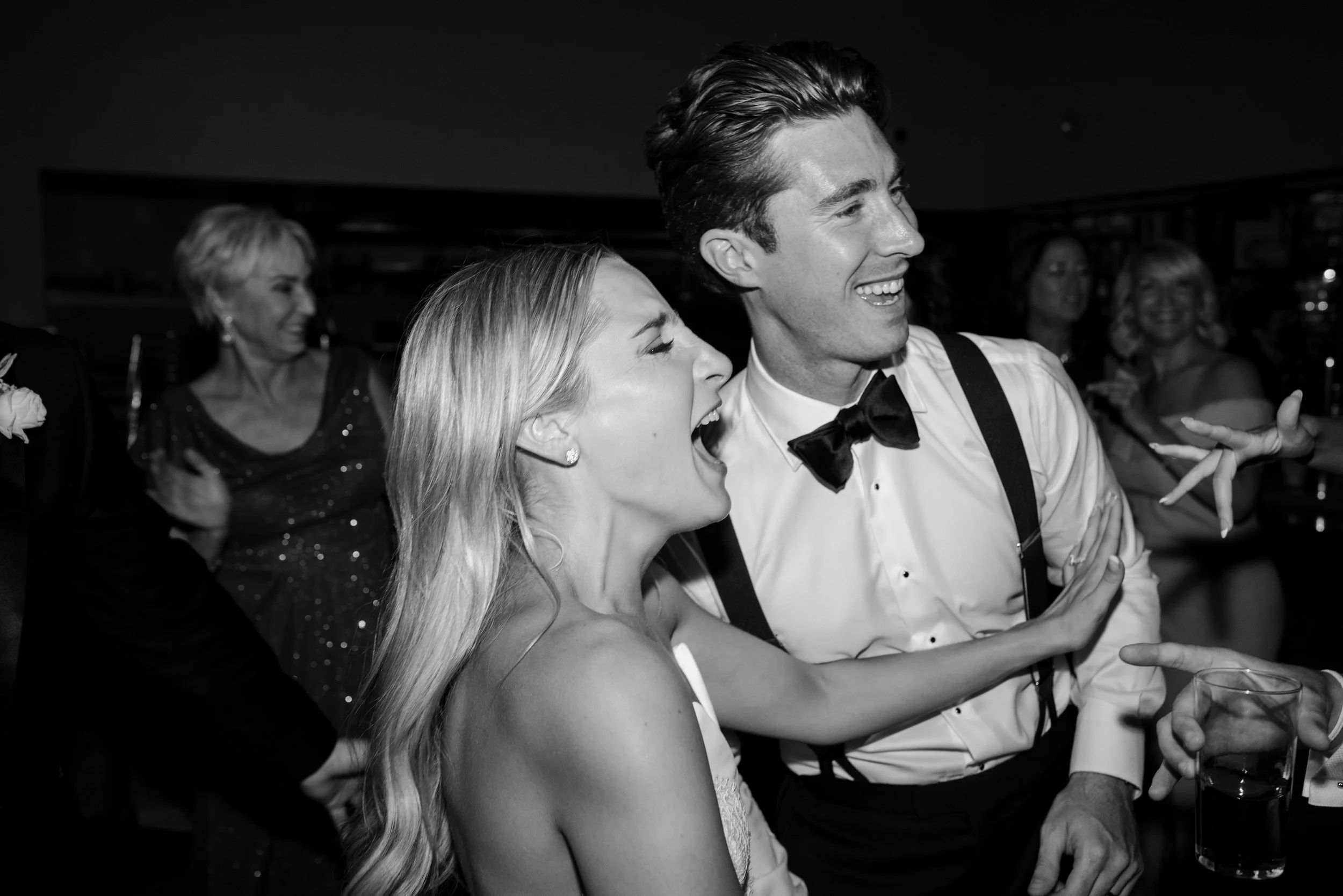 A black-and-white photo of a group of people at a party, with the focus on a smiling woman and a man dressed in formal attire, laughing and engaging with each other.