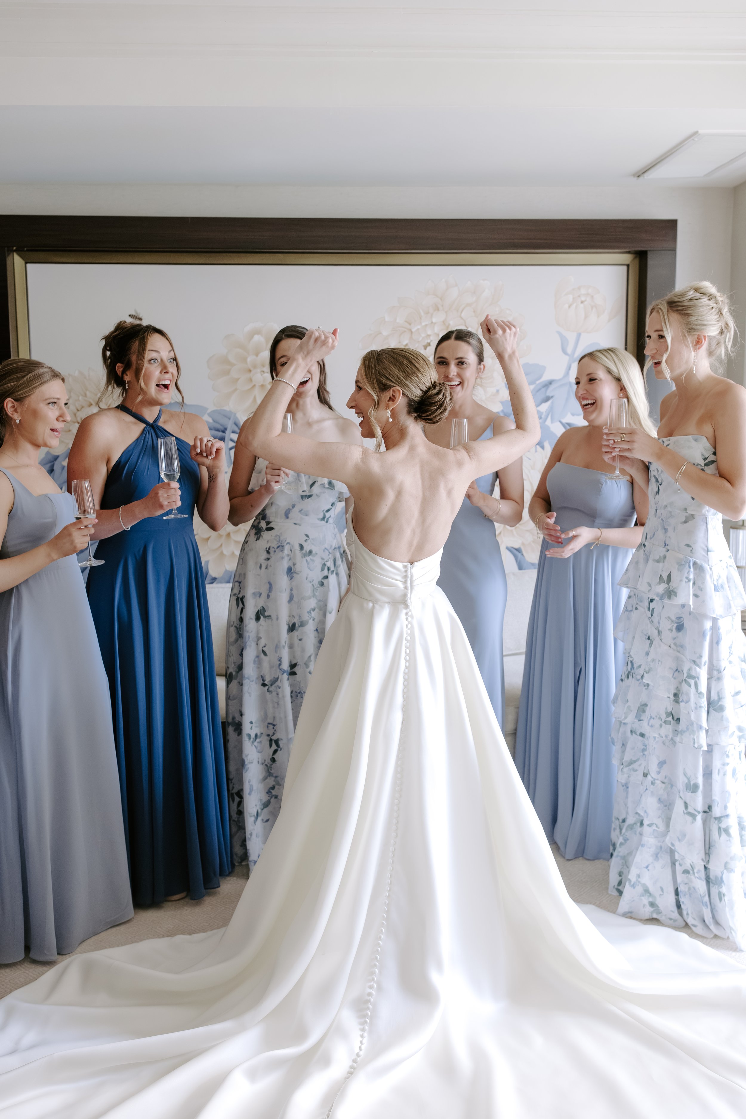 A bride surrounded by six bridesmaids celebrating in a room with a large floral mural backdrop, all holding champagne glasses and smiling.