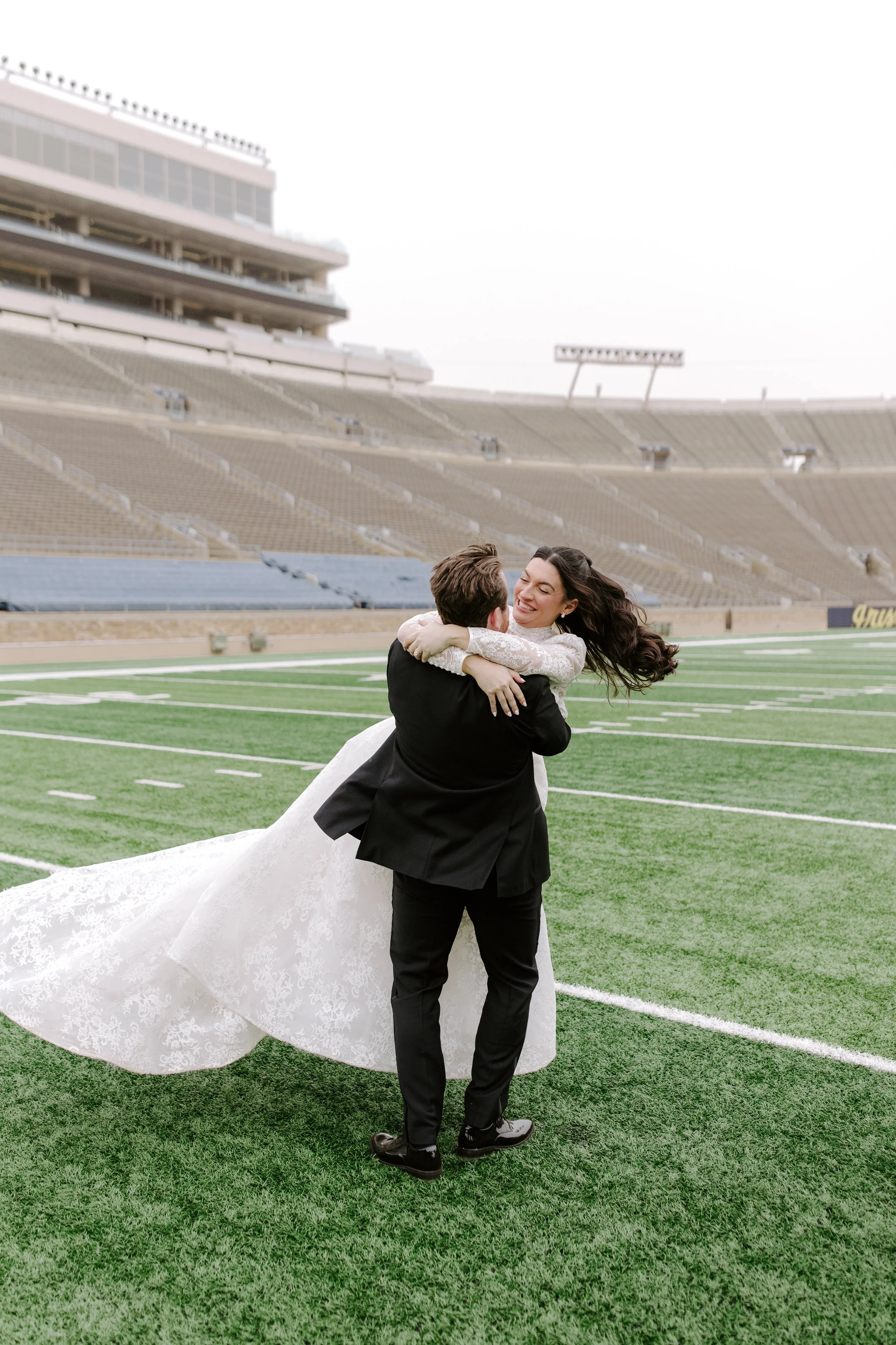 A bride and groom sharing a joyful hug on a football field in a stadium, with empty bleachers and an open sky in the background.