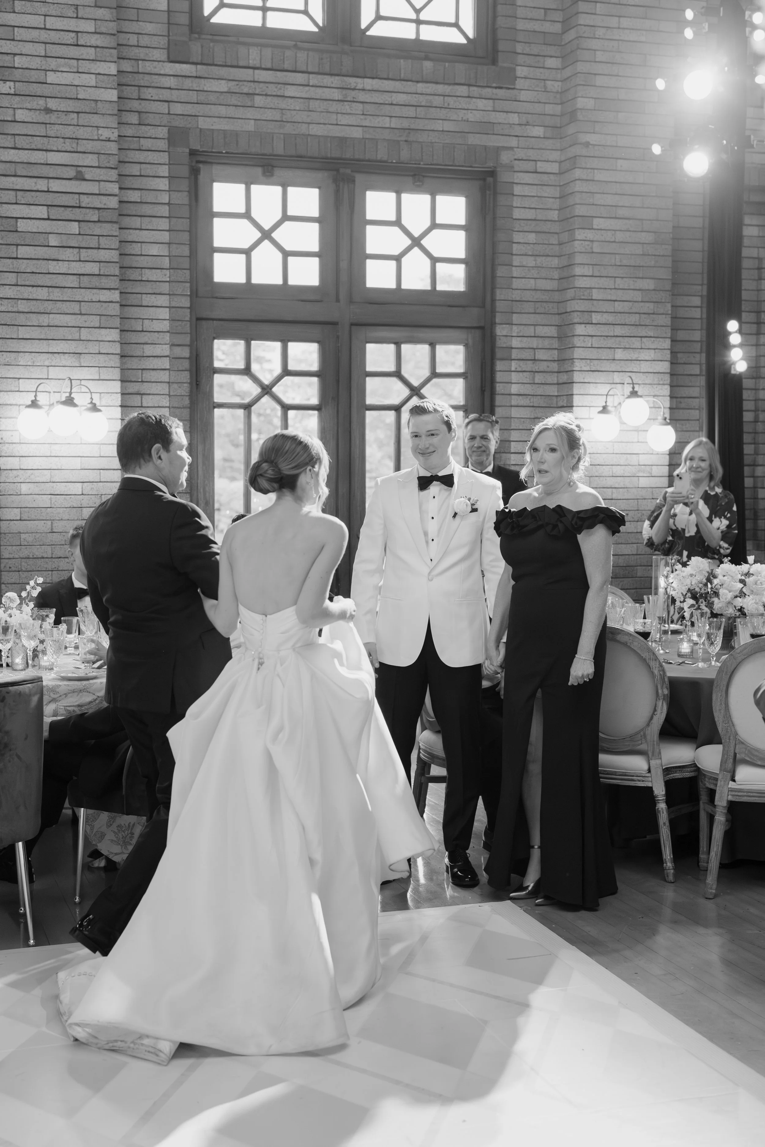 A wedding reception with a bride in a strapless gown and a groom in a tuxedo exchanging vows or having a moment, surrounded by family and friends in a decorated indoor venue with large windows and brick walls.
