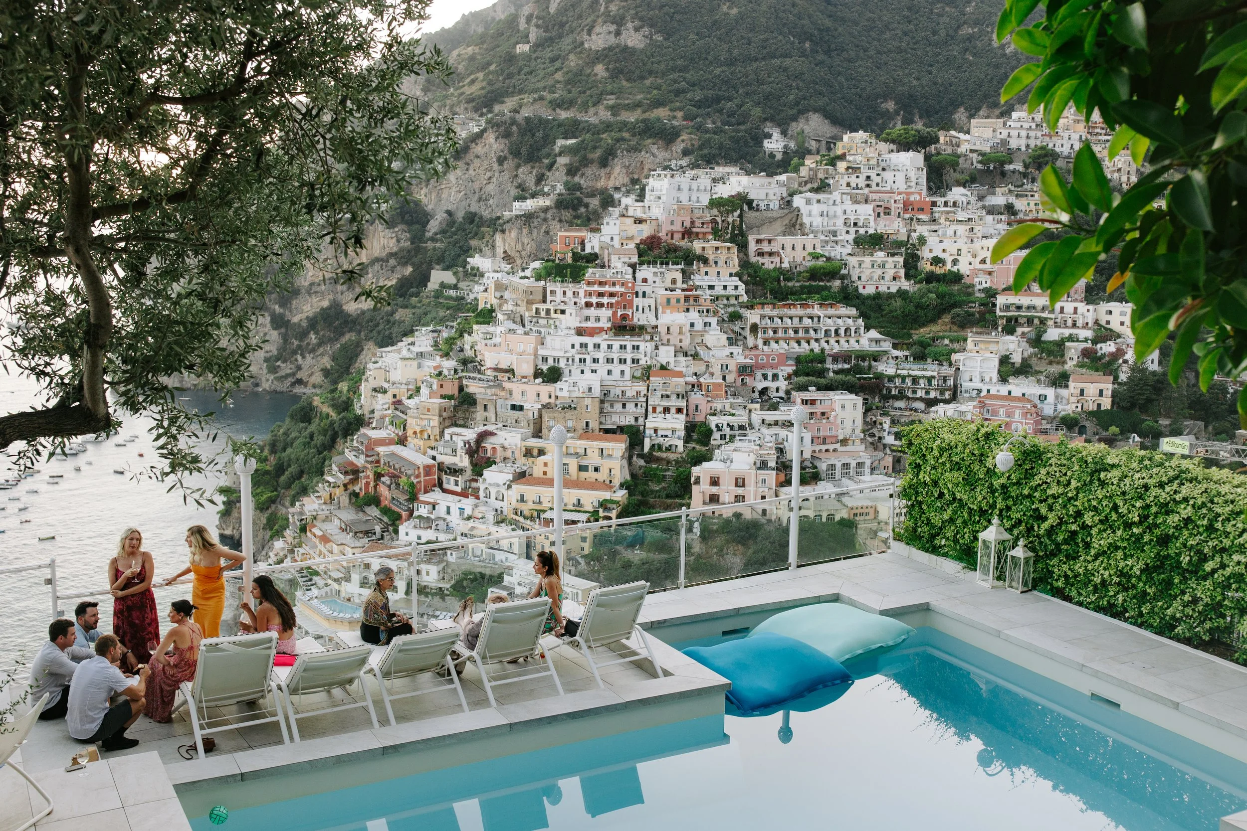 People gathering around a poolside patio with scenic hillside town in the background.