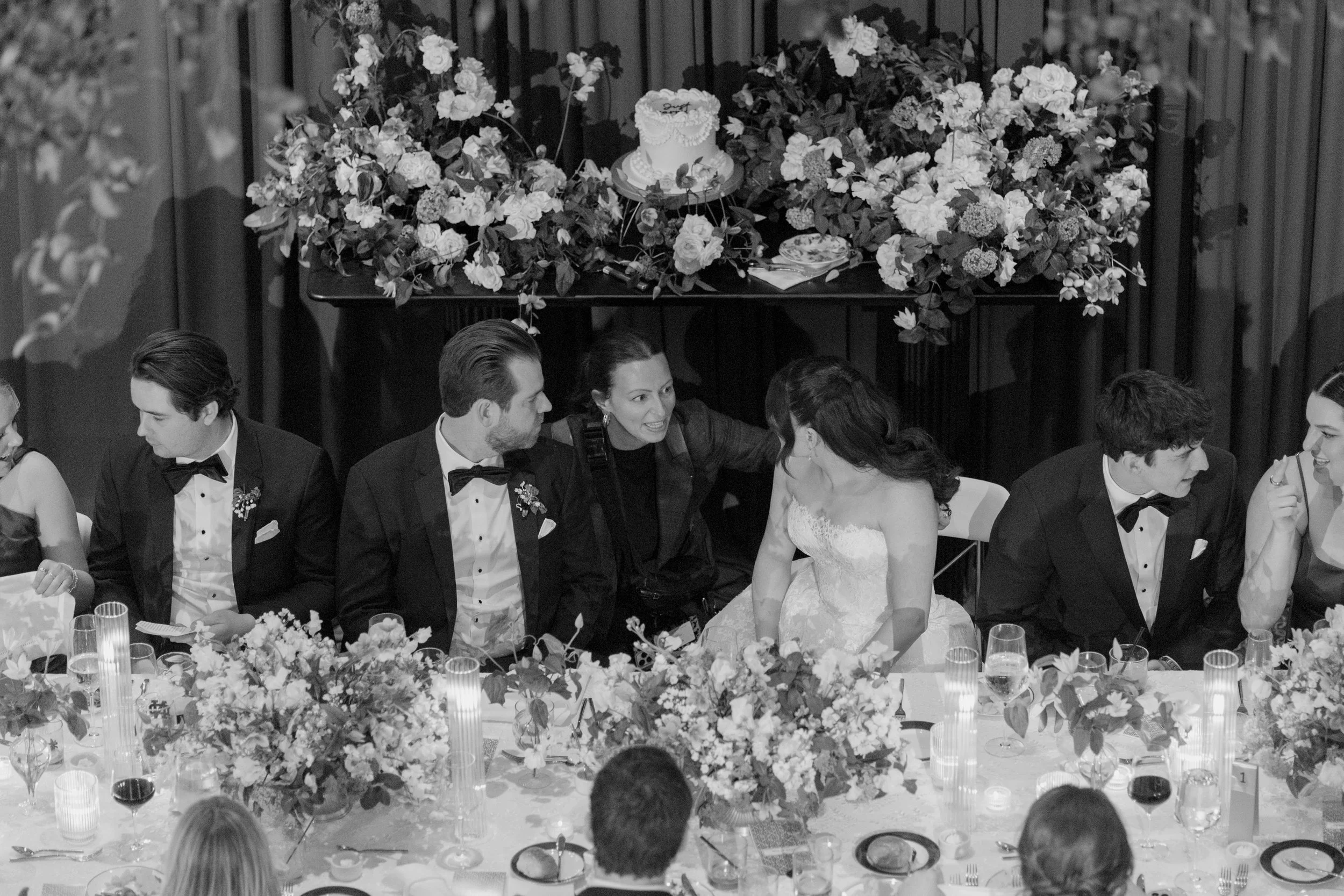 Black and white photo of a wedding reception with a long table decorated with flowers and candles. In the background, a wedding cake is displayed on a table surrounded by flower arrangements. Several couples are seated at the table, dressed in formal