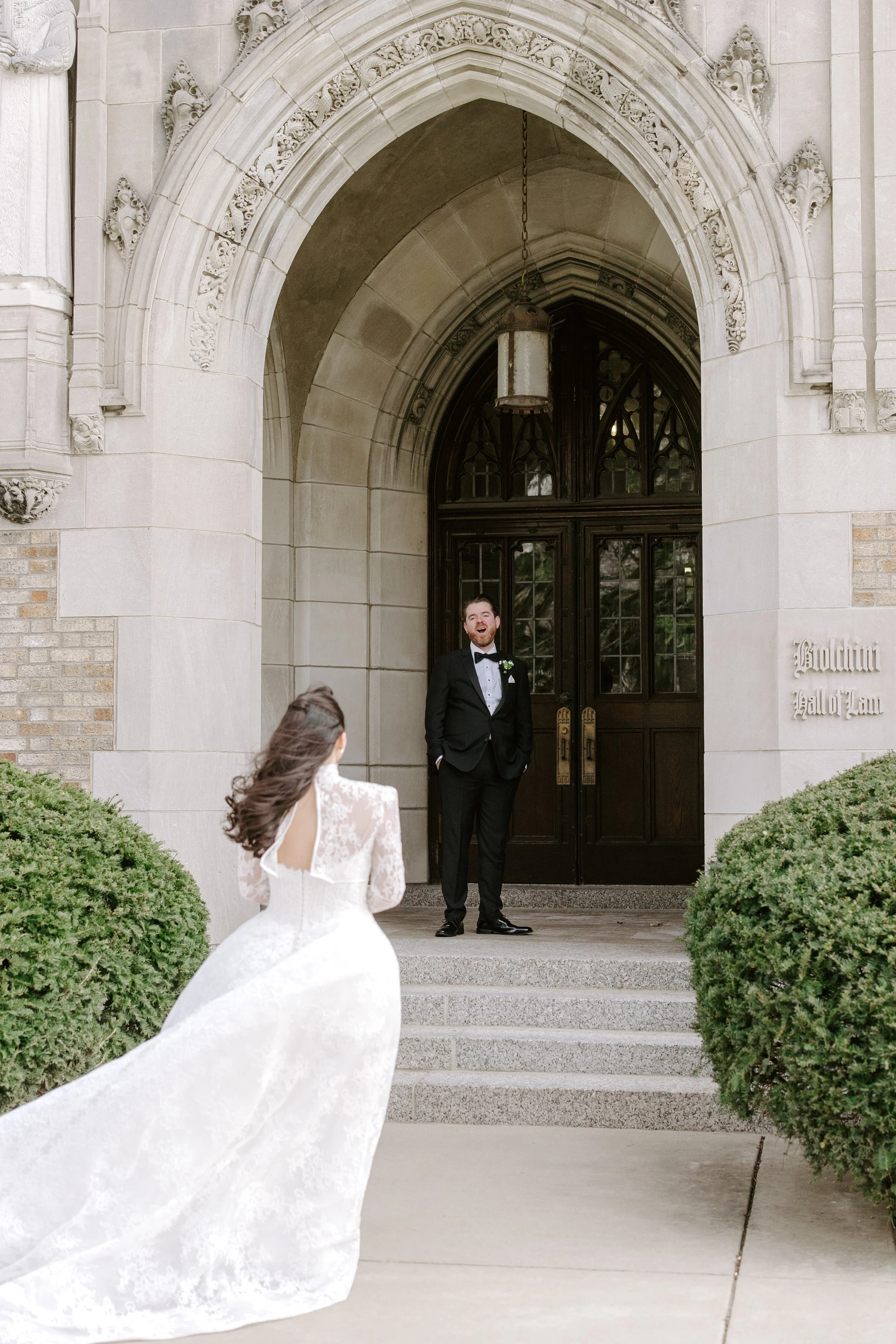 A wedding scene with a bride in a white wedding dress and a groom in a black tuxedo standing on the steps of a historic building. The bride's dress has a long train flowing behind her and she is facing the groom, who is smiling and looking at her.