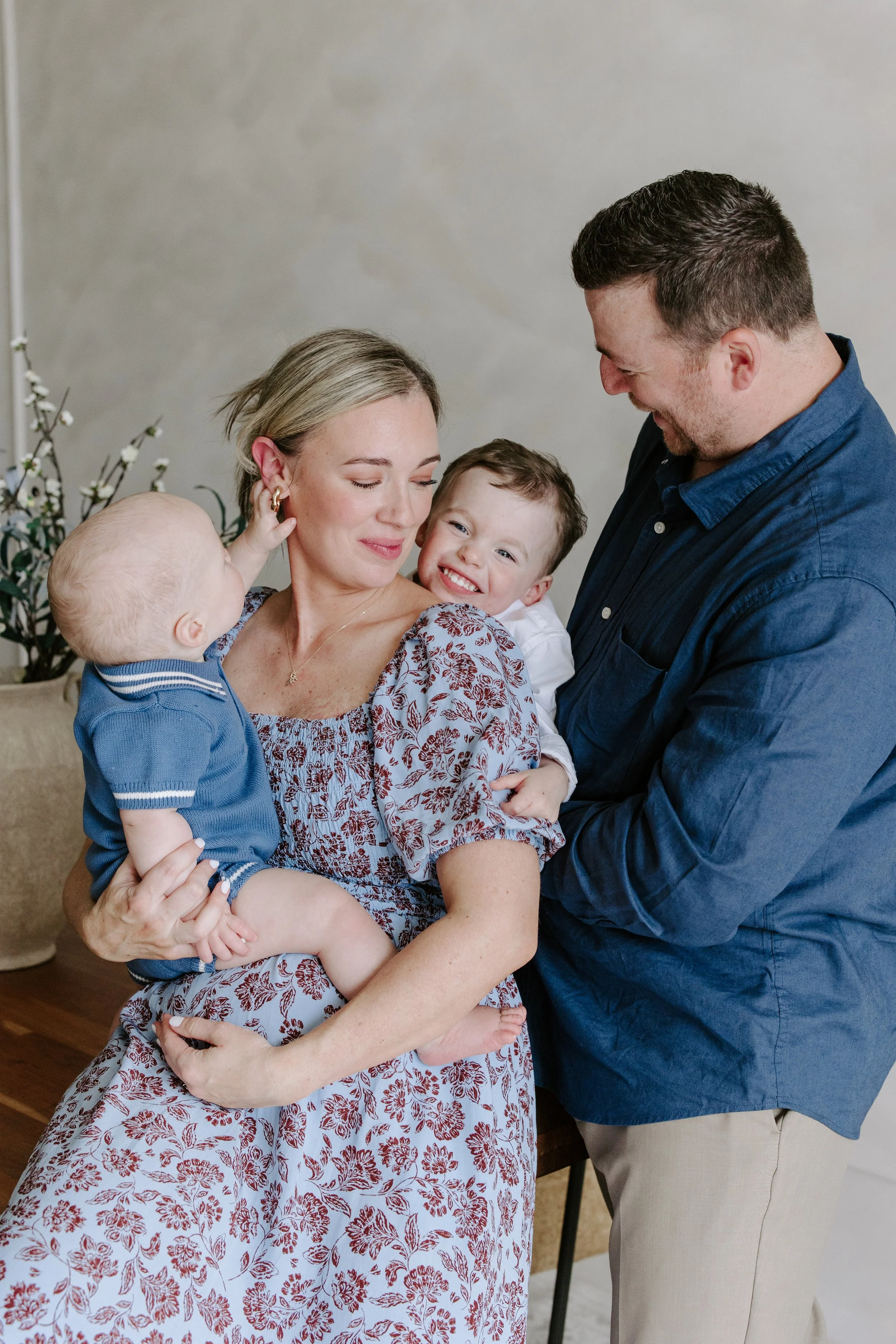 A family of four, including a woman with blonde hair, a man with short brown hair, a young boy, and a baby, sharing a joyful moment indoors.